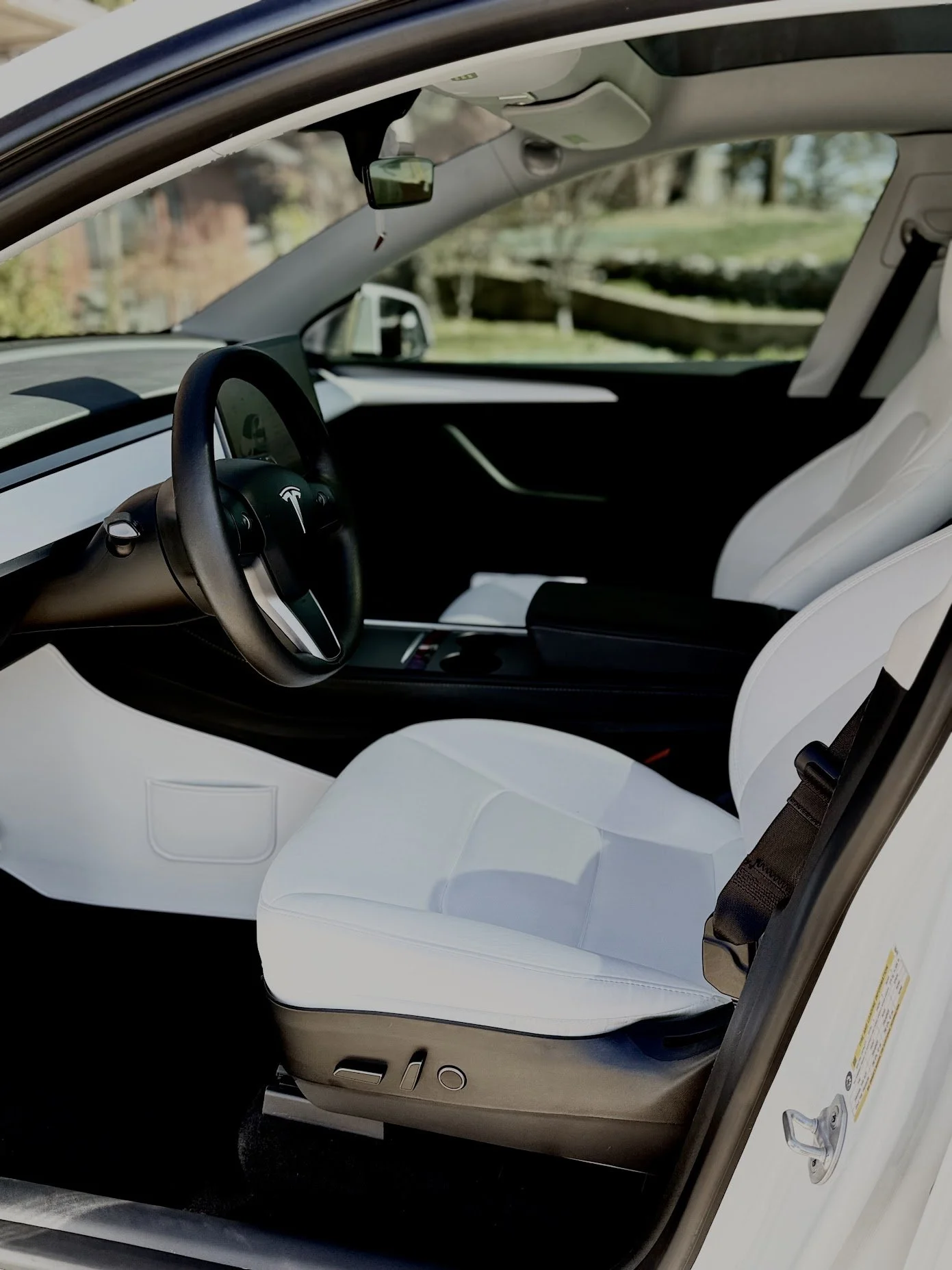 Interior of a Tesla Model S with white seats, steering wheel, and dashboard, taken from the driver's side door open, outdoor background with trees and a pond.