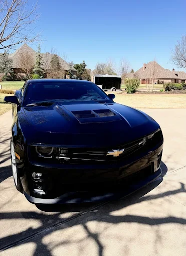 A black Chevrolet Camaro sports car parked in a driveway with a suburban neighborhood in the background.
