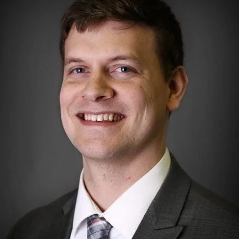 Headshot of a smiling man in a suit and tie against a dark background.