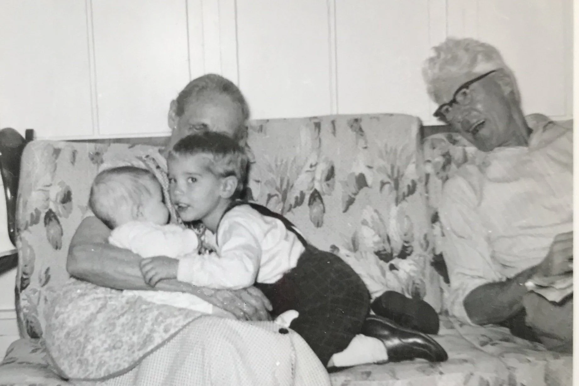 A black and white photo showing a woman sitting on a floral couch, holding a small dog, with a young girl hugging her and a man smiling and leaning in from the right, displaying the concept of Multigenerational Living.