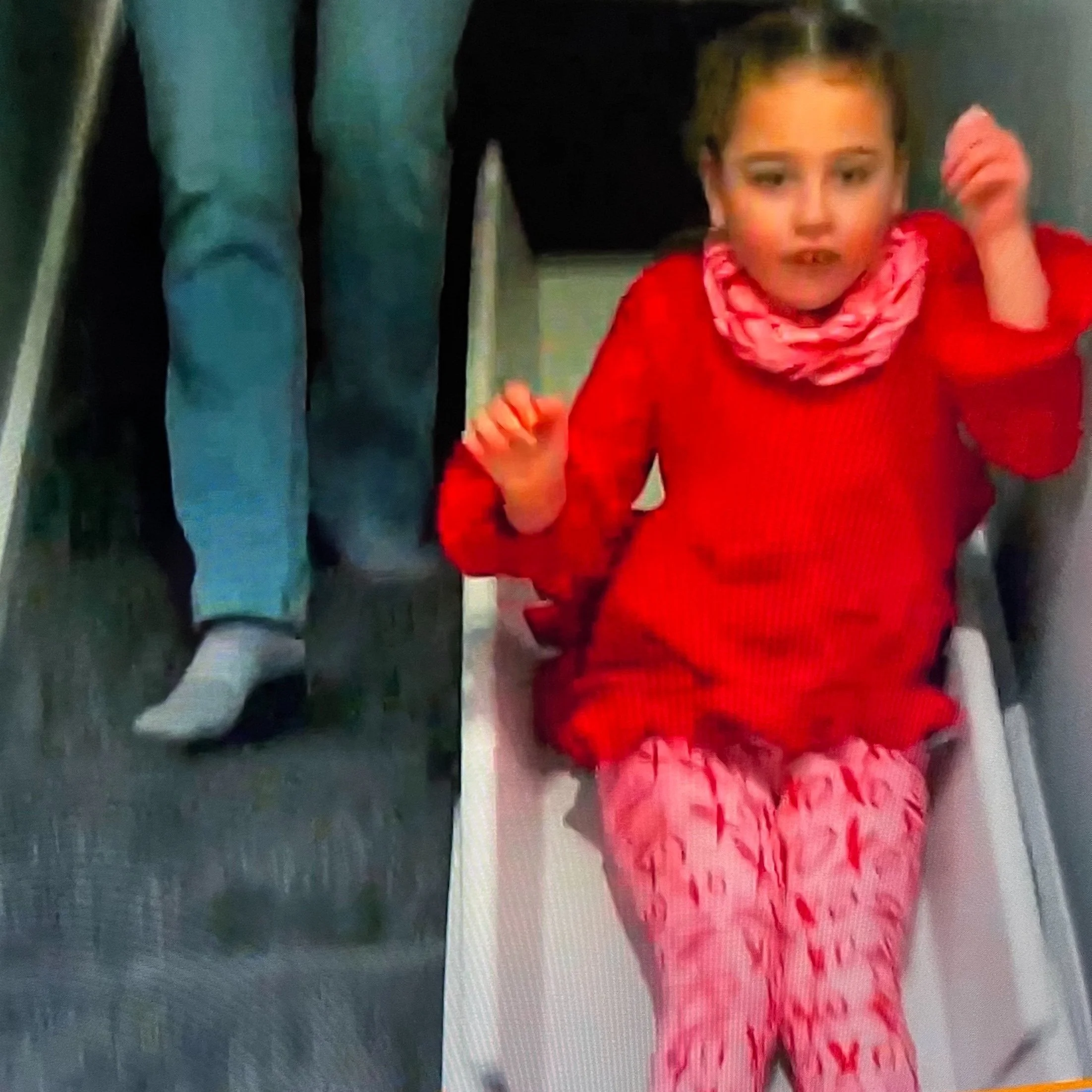 A young girl sitting on a plastic chair in a hallway, wearing a red top and pink pajama pants with a butterfly pattern, with two adults standing beside her.