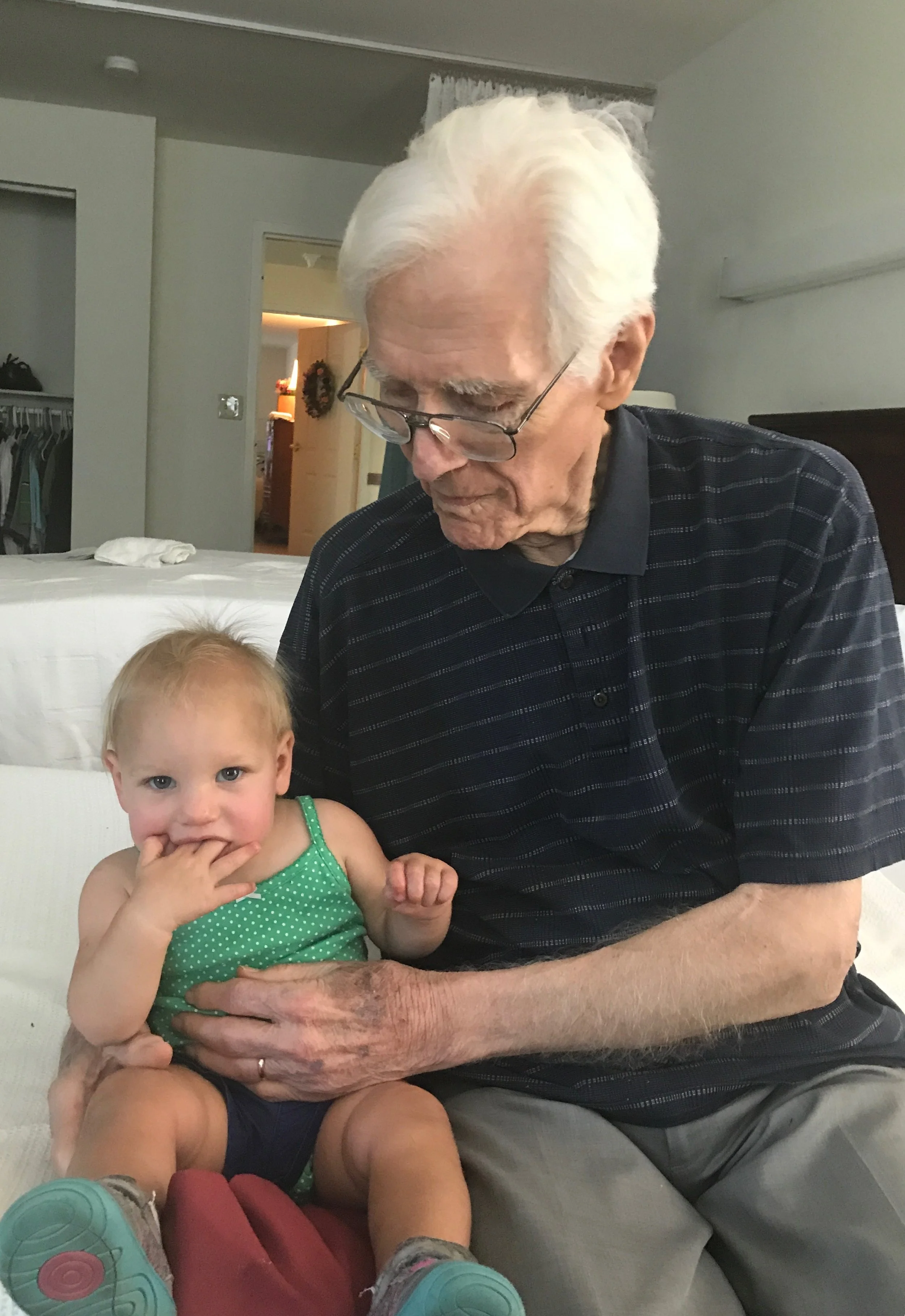 An elderly man with white hair and glasses sitting on a bed, holding a young girl with blonde hair in a green polka dot shirt, who is sitting on his lap and has her fingers in her mouth.