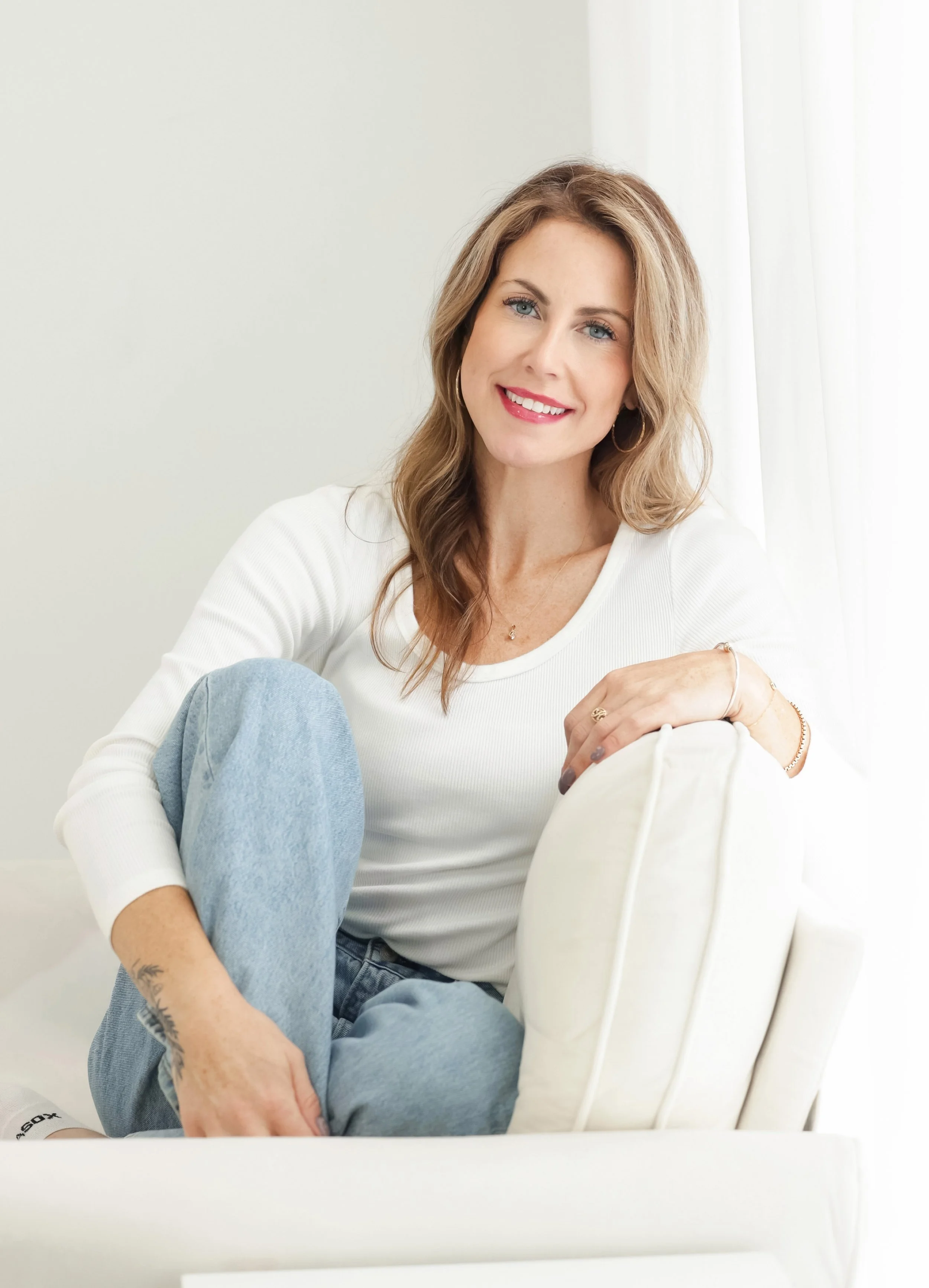 A woman with long wavy blonde hair, wearing a white long sleeve shirt and light blue jeans, sitting on a white chair. She has blue eyes, is smiling, and looking at the camera. The background is a light-colored wall with a white curtain.