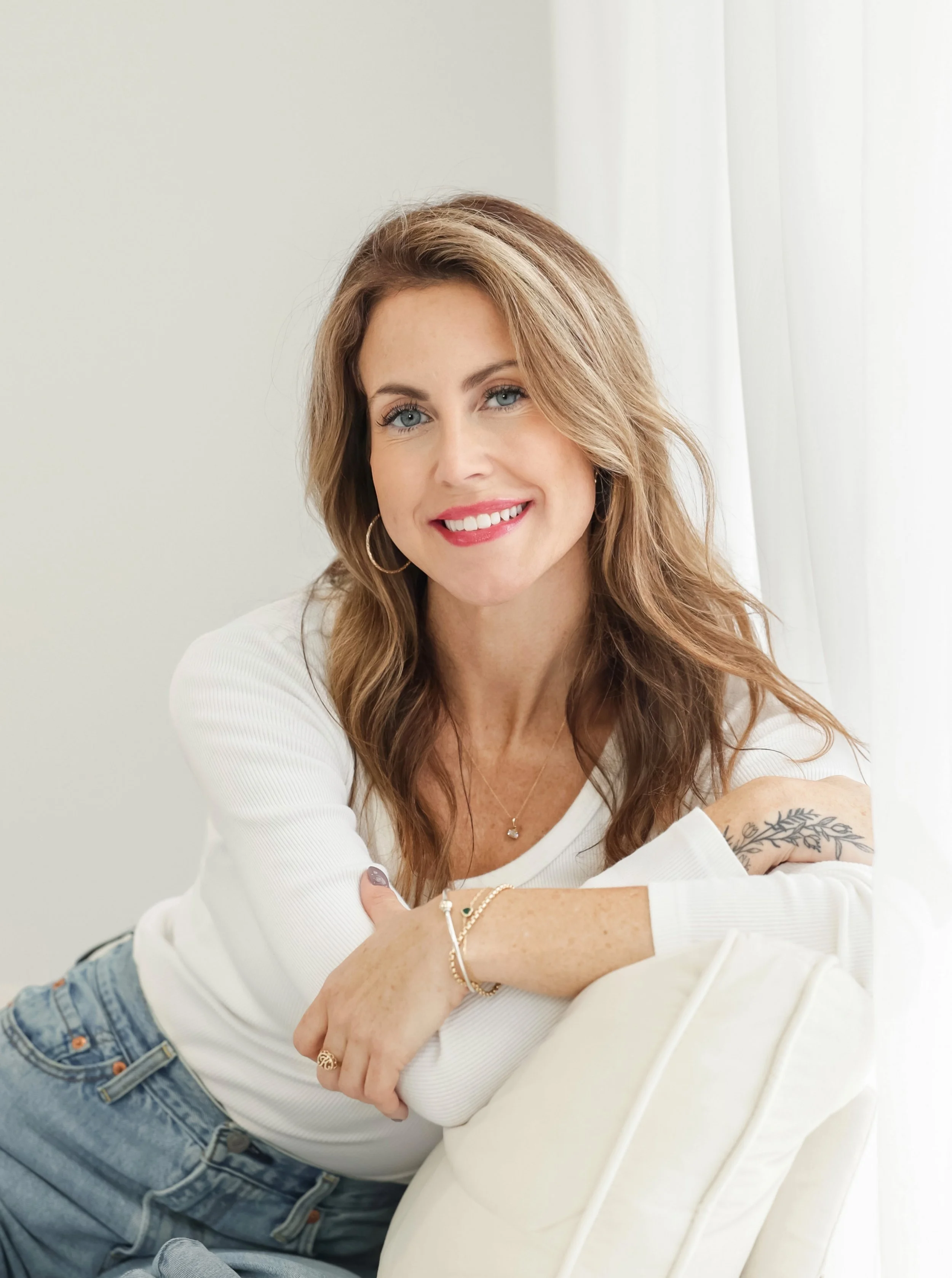 A woman with wavy, shoulder-length brown hair, wearing a white long-sleeve top and blue jeans, smiling and leaning on a white pillow in a bright room.