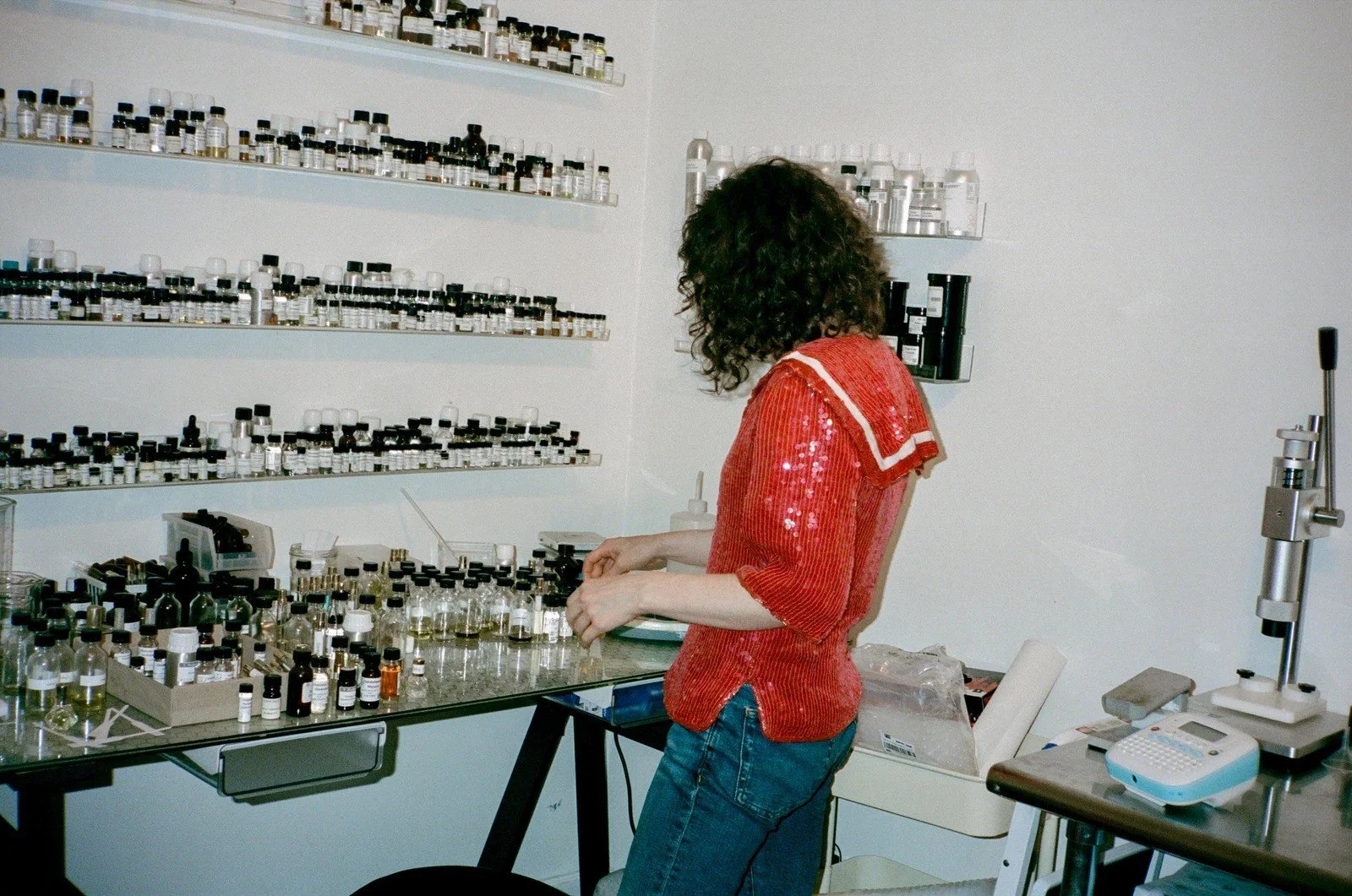 A person with curly hair wearing a red top and jeans working with small bottles and vials in a laboratory or apothecary setting, with shelves of bottles behind them.