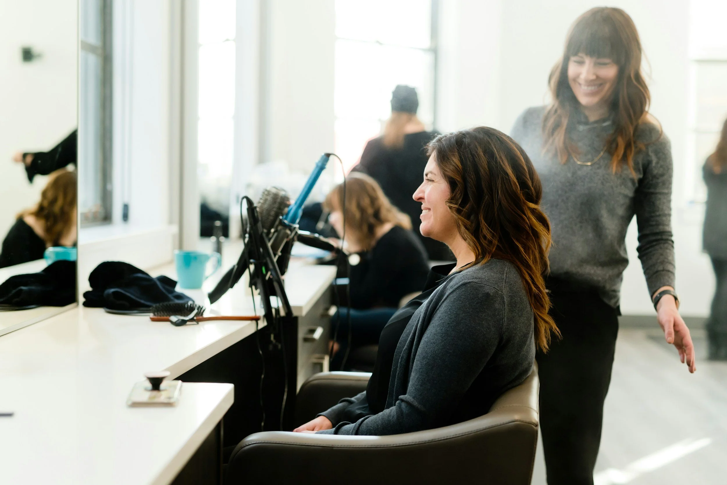 A woman sits in a salon chair getting her hair styled, smiling with her eyes closed, while a hairstylist stands behind her, smiling.