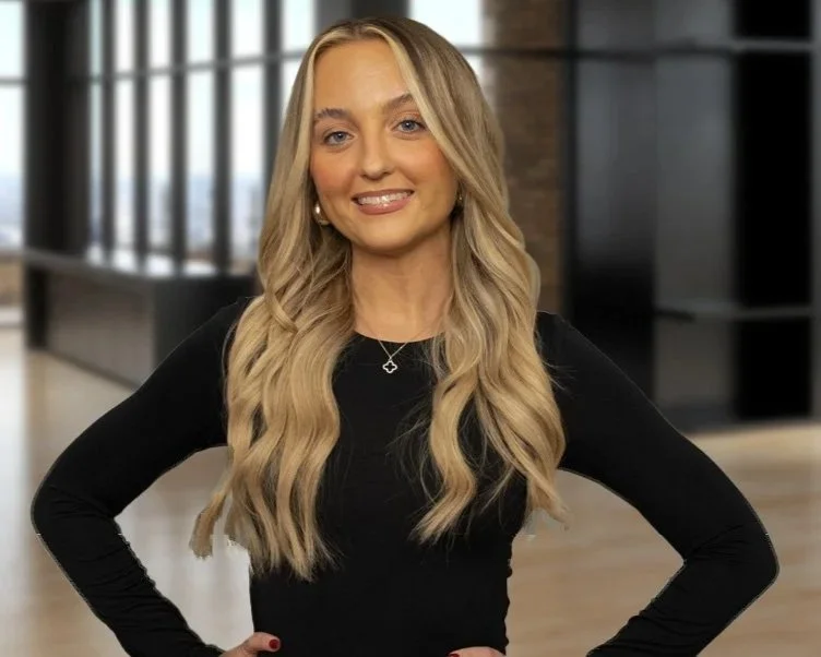 A woman with long, wavy blonde hair smiling in a modern office setting, wearing a black long-sleeve top and jewelry.