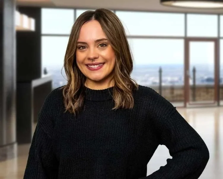 A smiling woman with shoulder-length wavy brown hair, wearing a black sweater, standing in a modern room with large windows and a city view in the background.