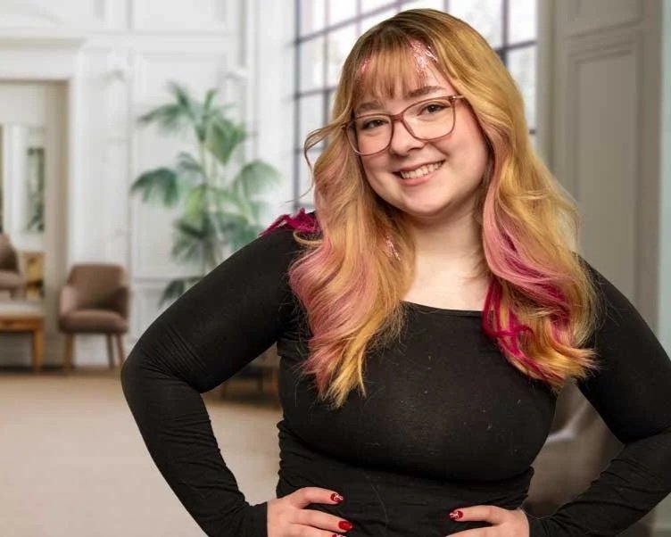A smiling woman with glasses and colorful hair standing in a bright, modern room with large windows and green plants.