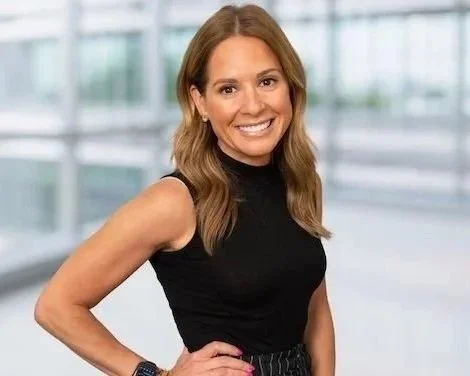 A woman smiling in an indoor office setting with large windows.