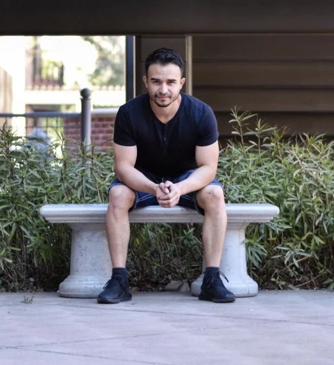 Man sitting on a stone bench outdoors, wearing a black t-shirt and shorts, with greenery in the background.
