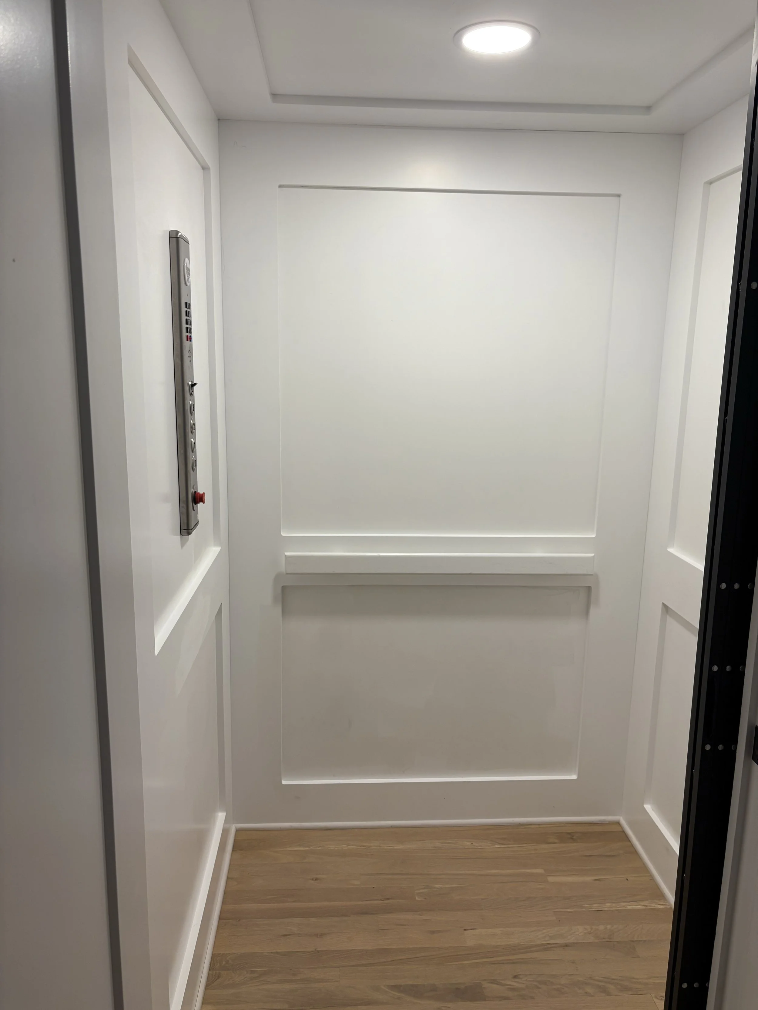 Inside an empty elevator with white walls, a wooden floor, and a control panel on the left wall containing buttons and an emergency button.