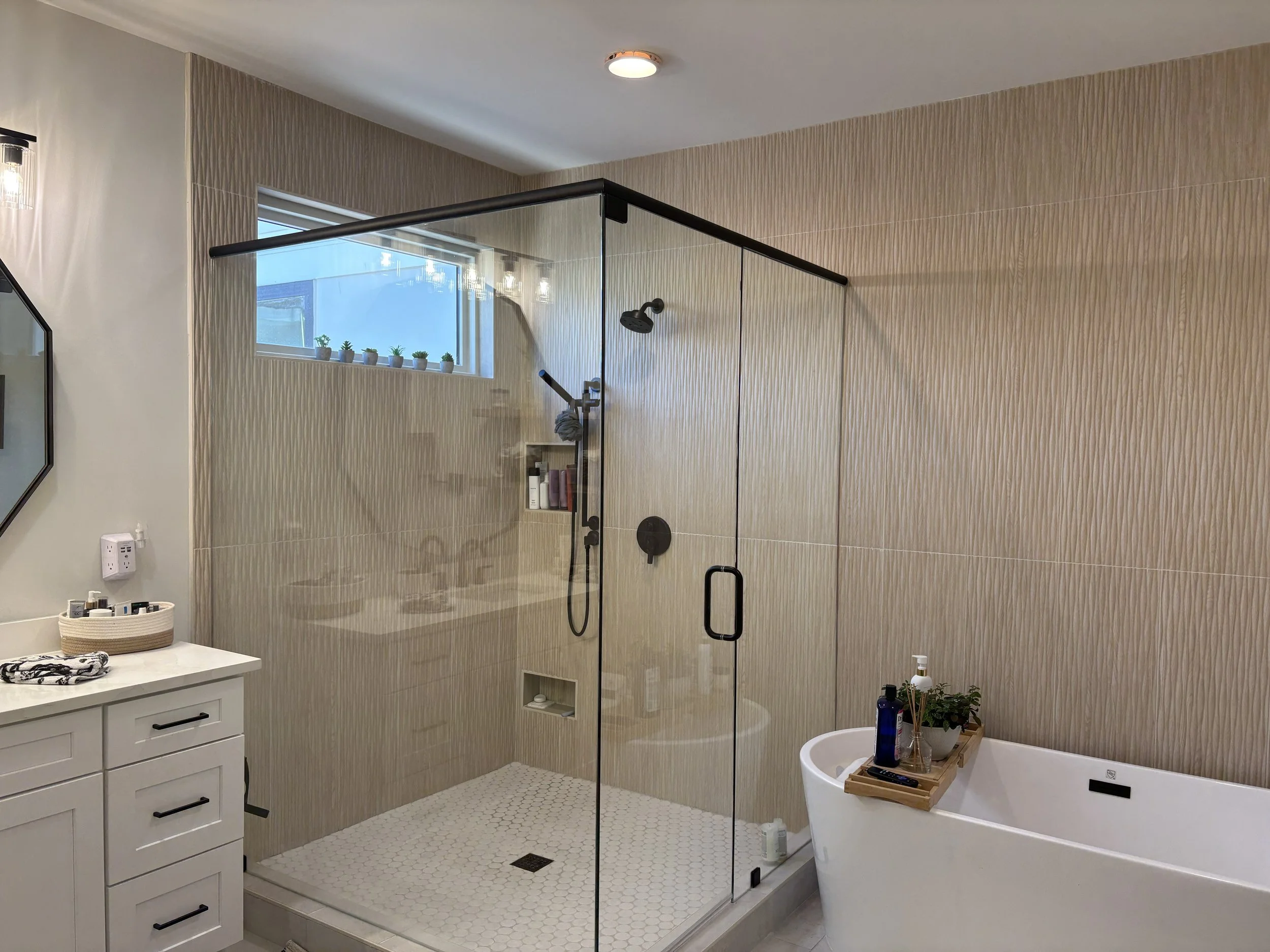 A modern bathroom featuring a glass shower enclosure with a black frame, beige textured wall tiles, a small window with potted plants, a white vanity with black handles, a wall-mounted mirror, a bathtub with a wooden tray holding toiletries and a pla