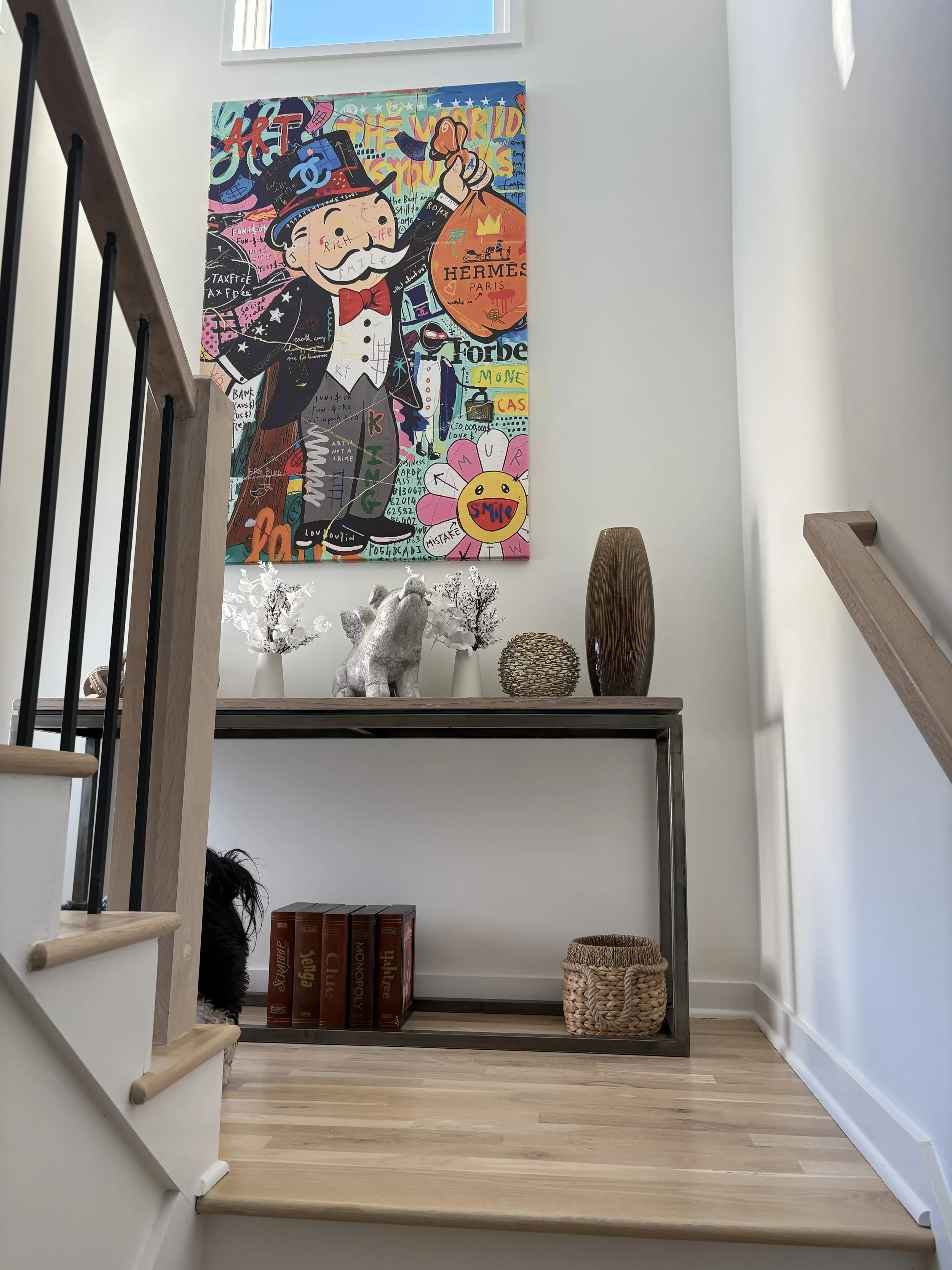 Interior view of a staircase landing with a colorful artwork hanging on the white wall, featuring a cartoon character in a top hat with a mustache, surrounded by various doodles and text. On the table below the artwork, there are decorative items inc
