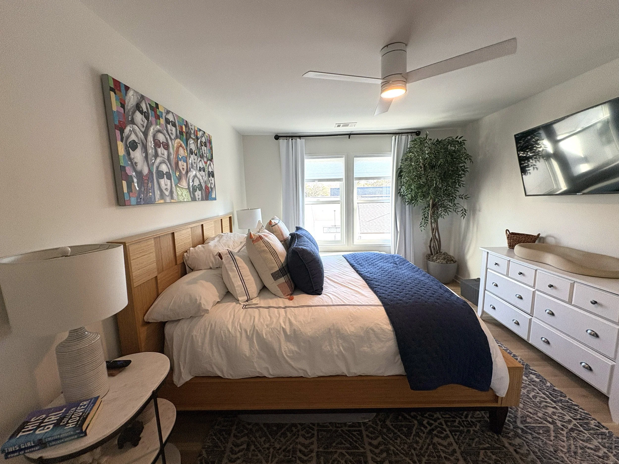 A bedroom with a wooden bed frame, white bedding with multiple pillows, a navy blue throw, white nightstand with a lamp and books, a large window with white curtains, a tall potted plant, a white dresser, wall-mounted TV, and colorful artwork of peop