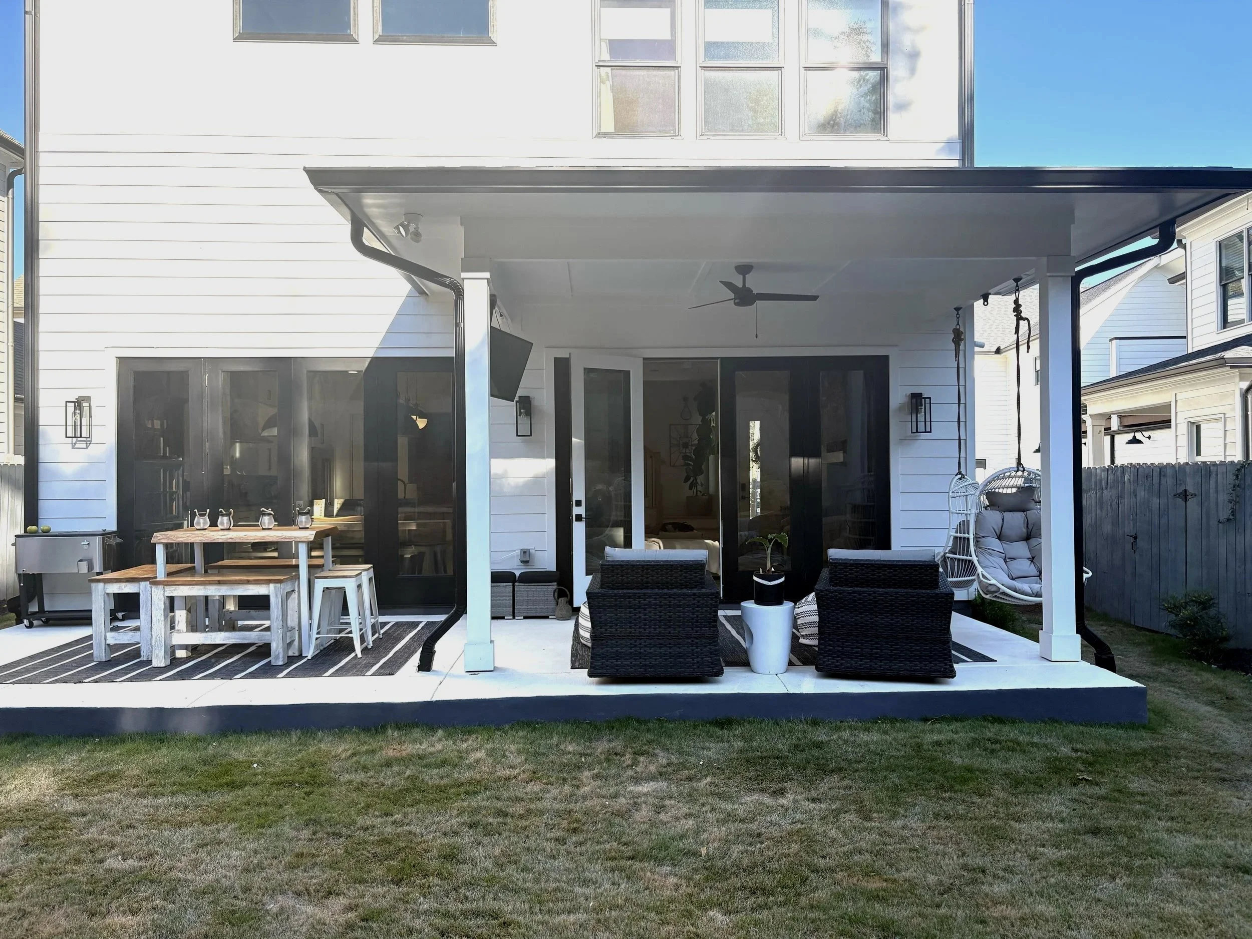Backyard patio with black and white furniture, hanging swing chair, ceiling fan, TV, and sliding glass doors, with lawn in foreground and neighboring houses visible.