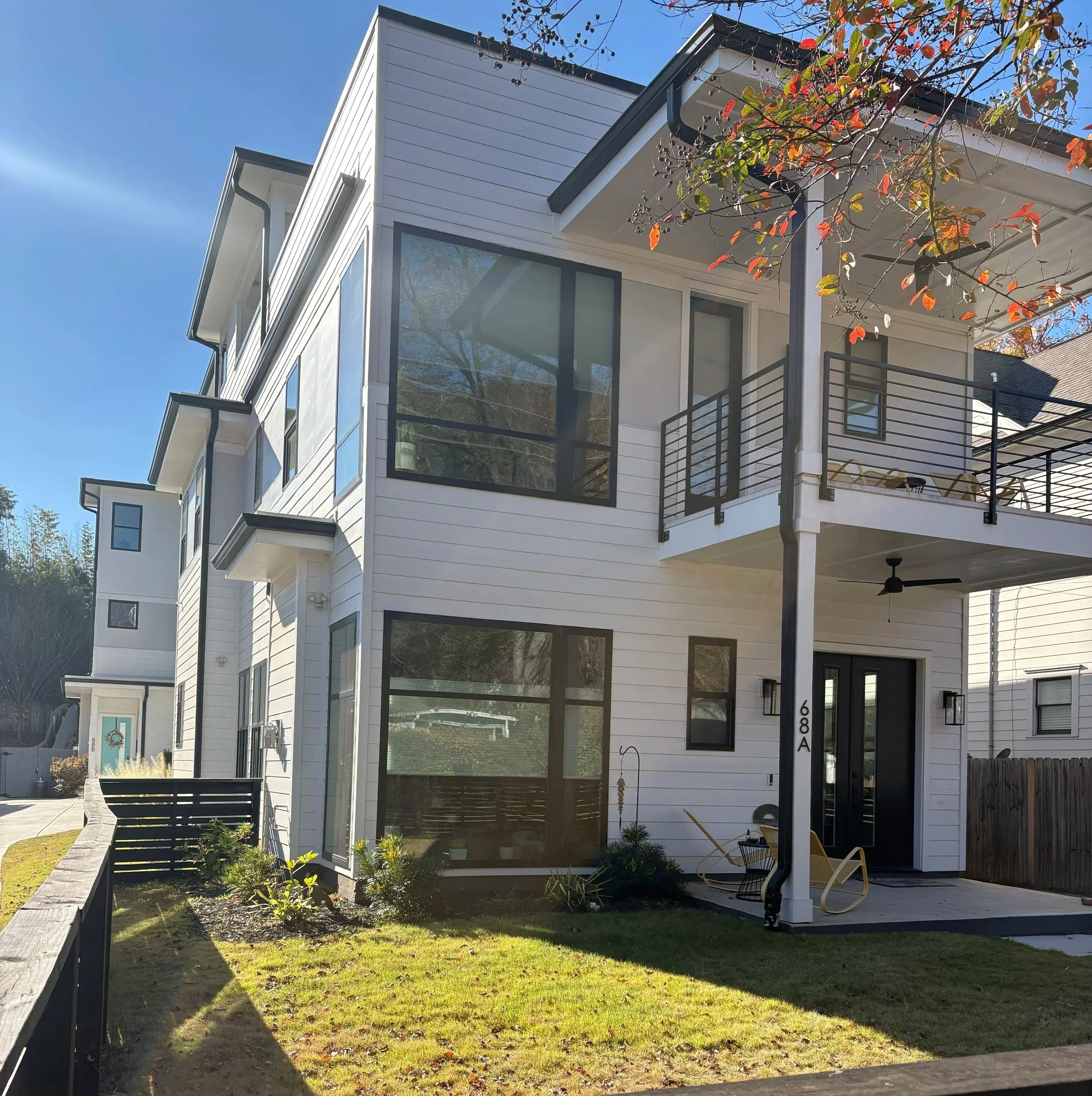 Modern white multi-story house with large windows, a balcony, and a porch with chairs and a ceiling fan, under a clear blue sky.