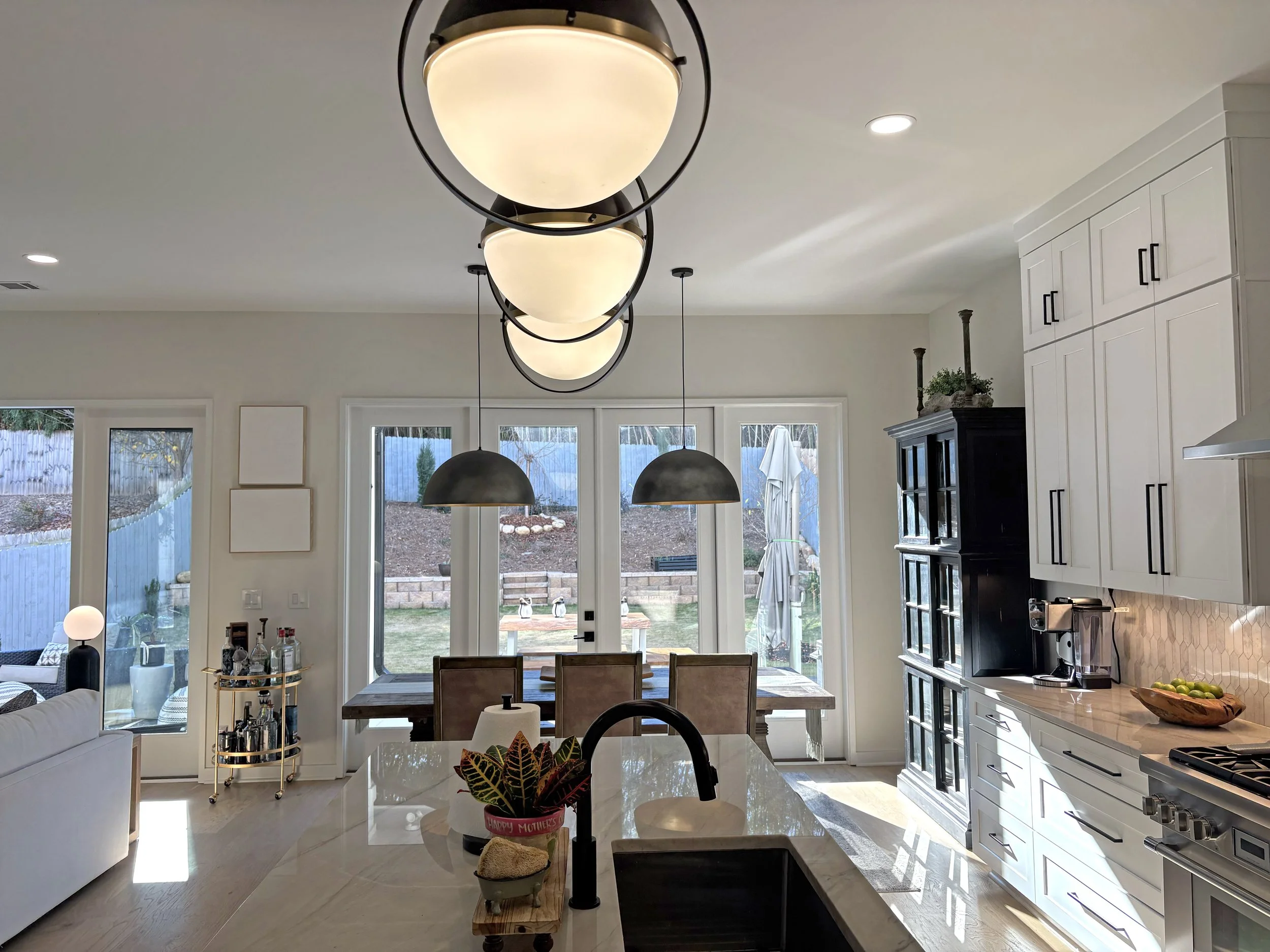 Modern kitchen with white cabinets, black hardware, a marble island, and a dining area with large windows and sliding glass doors leading to a backyard.