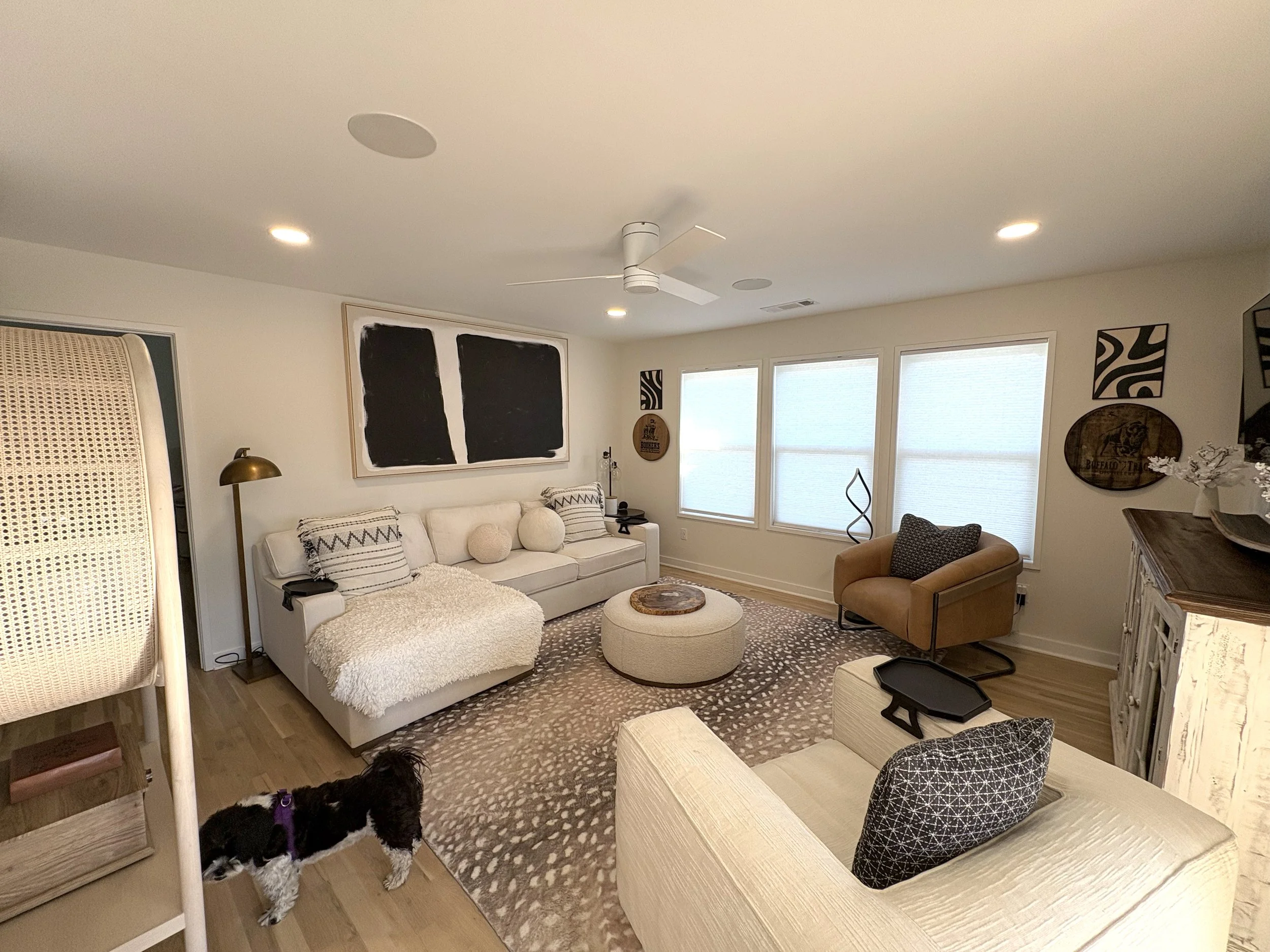 Living room with white sofa, beige armchair, cowhide rug, black and white artwork, and a small black dog with a purple harness.