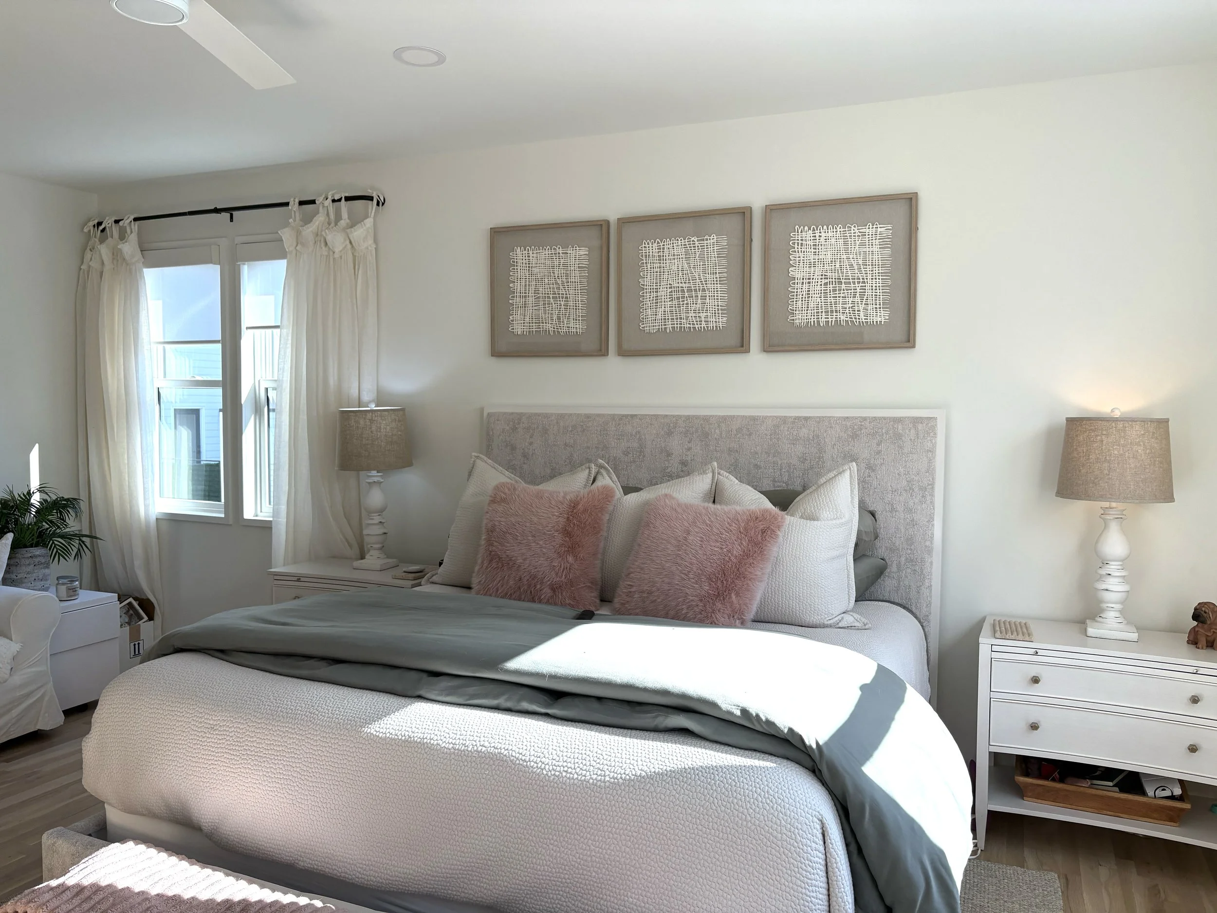 Bright bedroom with a large bed, light-colored quilt, pink accent pillows, and beige headboard. Sunlight streams through a window with white curtains. Two white nightstands with lamps, decorative objects, and a dog figurine, with three abstract frame