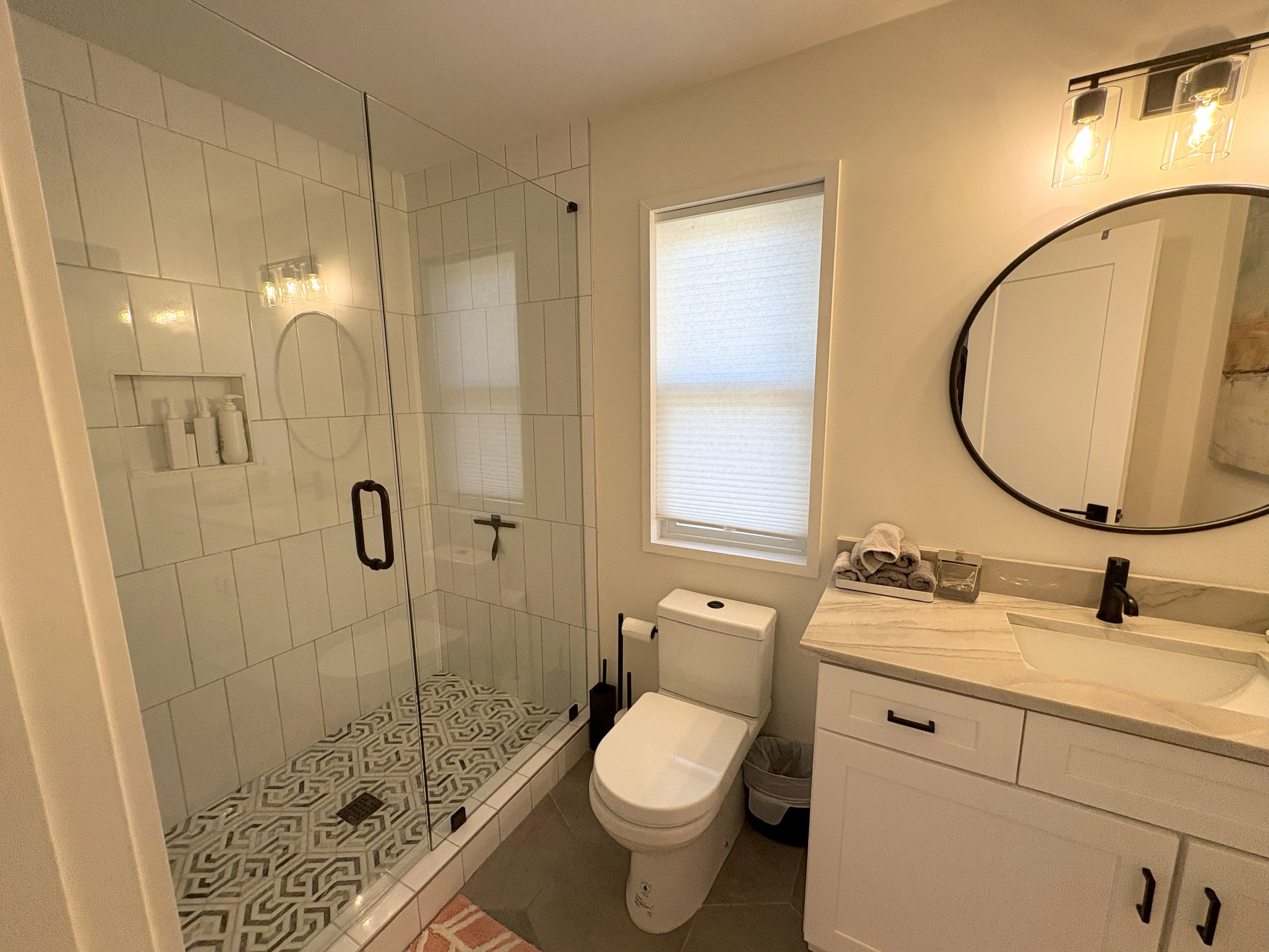 A modern bathroom with a glass-enclosed shower, a white toilet, and a white vanity with a marbled countertop and an oval mirror. There are folded towels on the countertop and a window with a roller shade. The lighting fixture has three hanging bulbs.