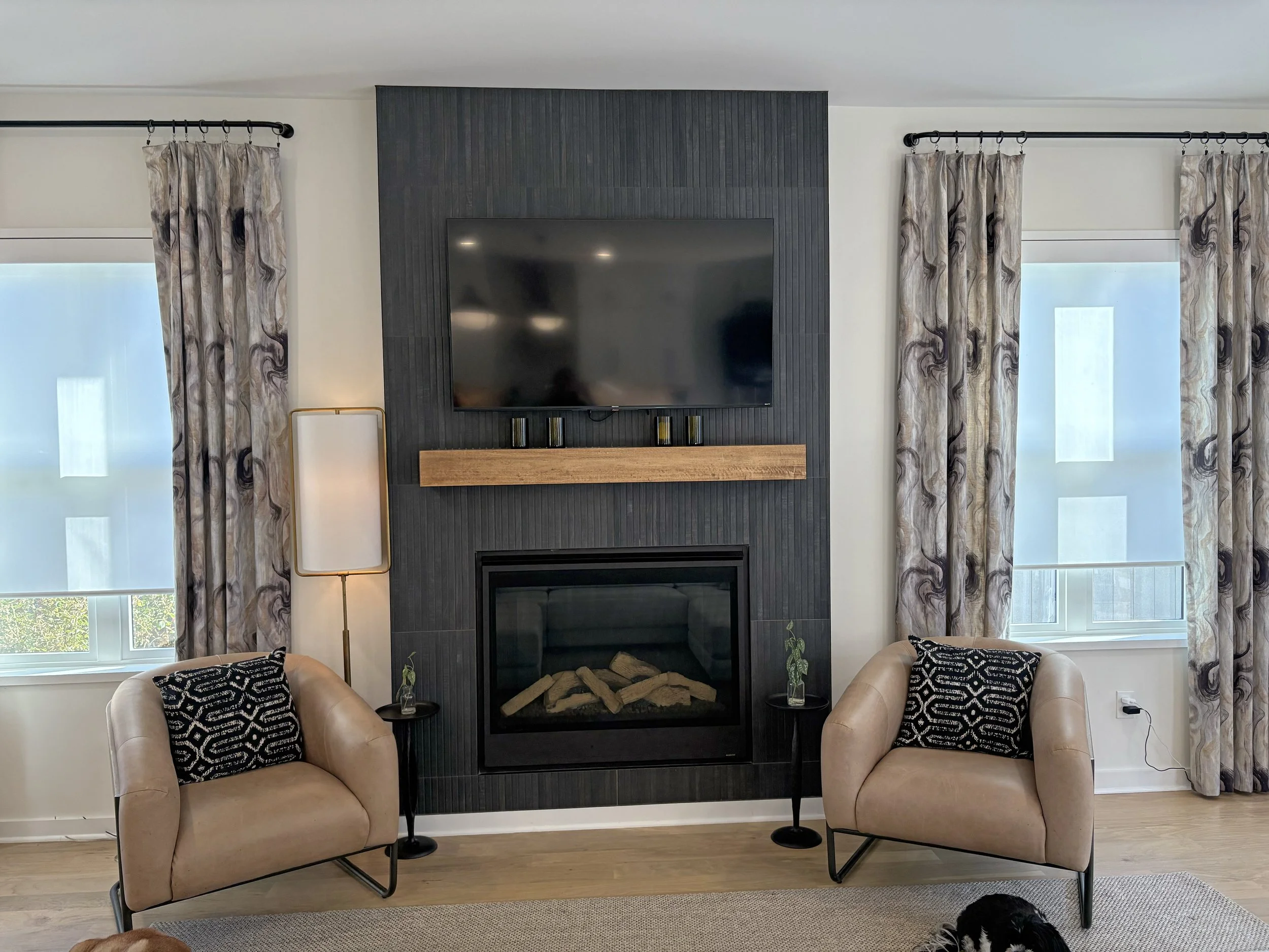 Living room with fireplace, two beige armchairs with black and white patterned pillows, two small side tables with plants, large window with patterned curtains, wall-mounted flat-screen TV, black accent wall, and wooden mantel above fireplace.