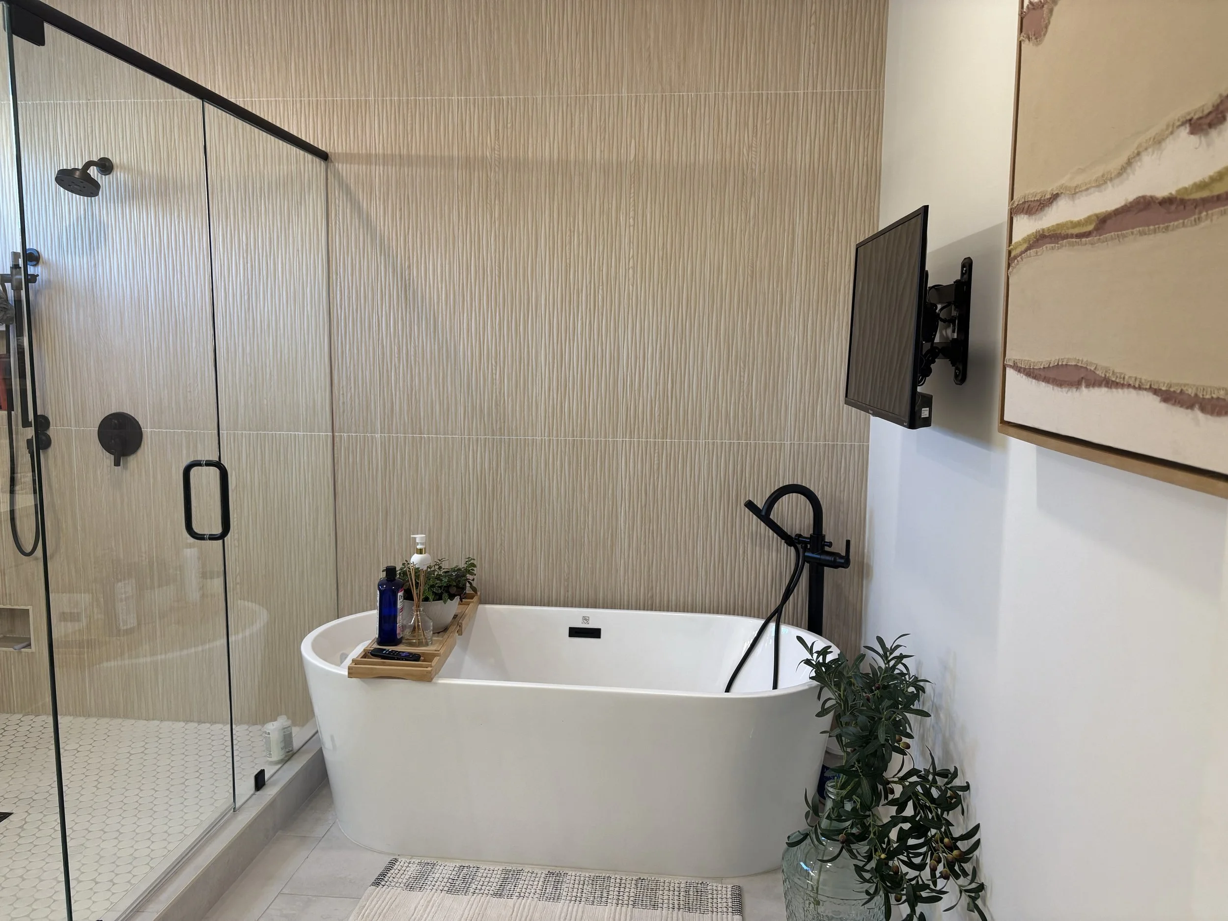 Modern bathroom with a free-standing bathtub, a walk-in shower with glass doors, a wall-mounted TV, plants, and artwork.