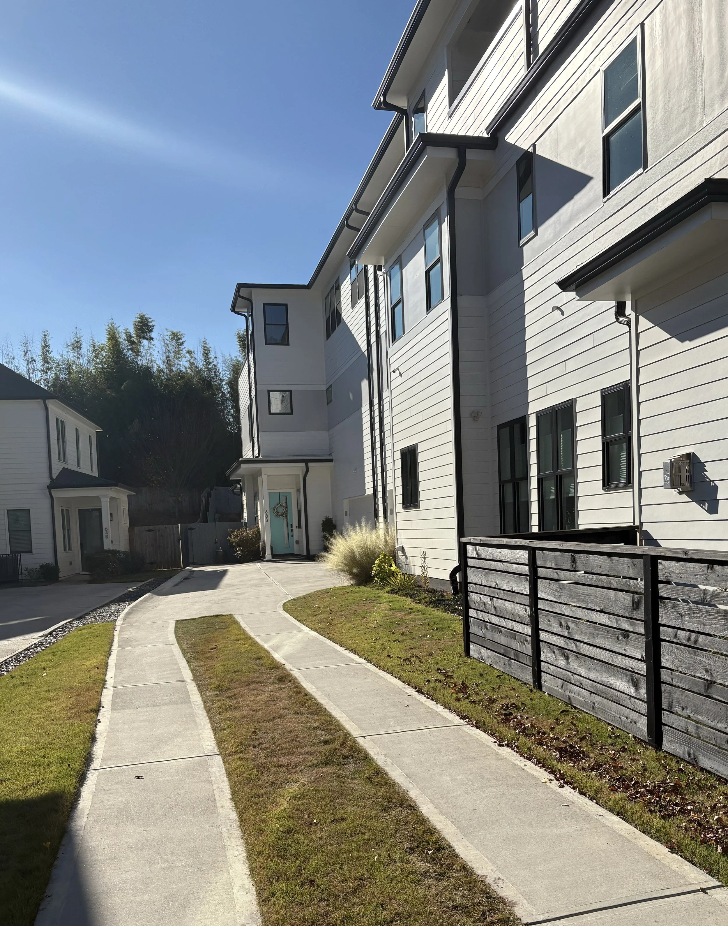 Modern white multi-story residential buildings with black window frames, a concrete walkway, and small patches of grass, under a clear blue sky.