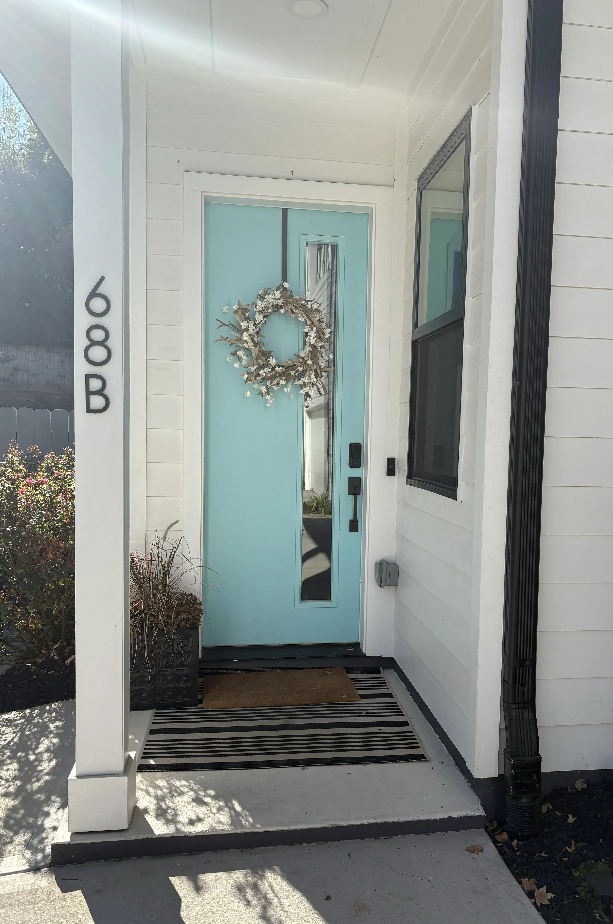Front porch of a house with a light blue door decorated with a white wreath of flowers and leaves, a black and white striped doormat, and the house number 968B displayed vertically on the left.