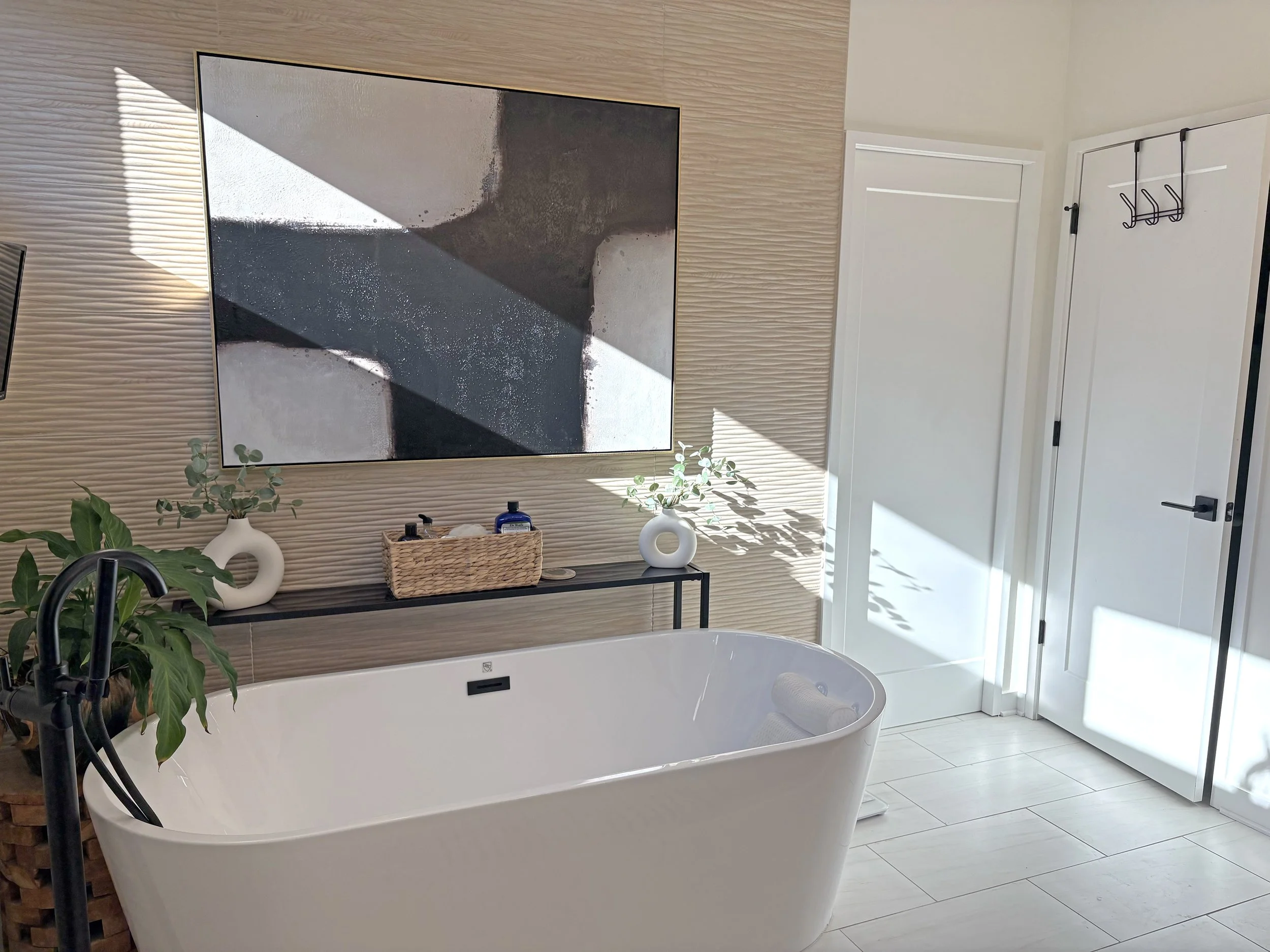Modern bathroom with a freestanding bathtub, two white vases with greenery, a small basket with toiletries, and a large abstract painting on a textured beige wall. Sunlight creates shadows on the wall and floor.