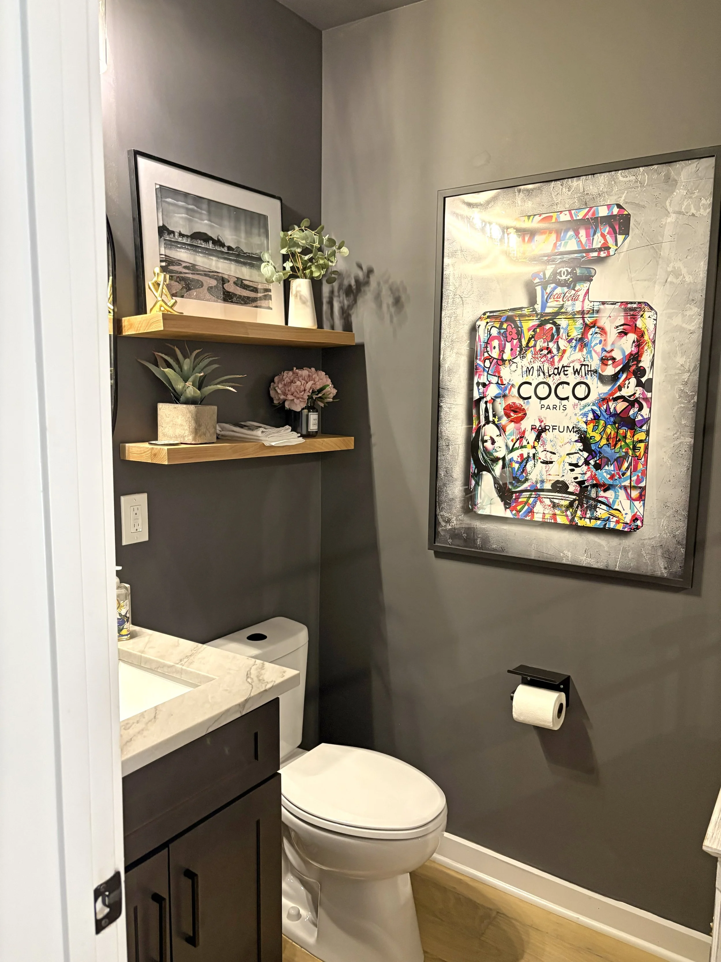 A small bathroom with a white toilet, gray walls, and wooden shelves holding decorative items. There is a large framed colorful pop art print of a perfume bottle on the wall and a roll of toilet paper on a holder.