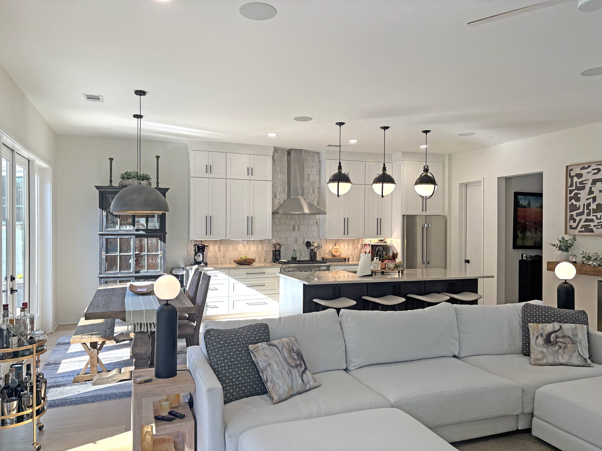 Open-concept living room and kitchen with white cabinetry, a large island with barstools, pendant lights, a dining table with chairs, a white sectional sofa, and decorative items.