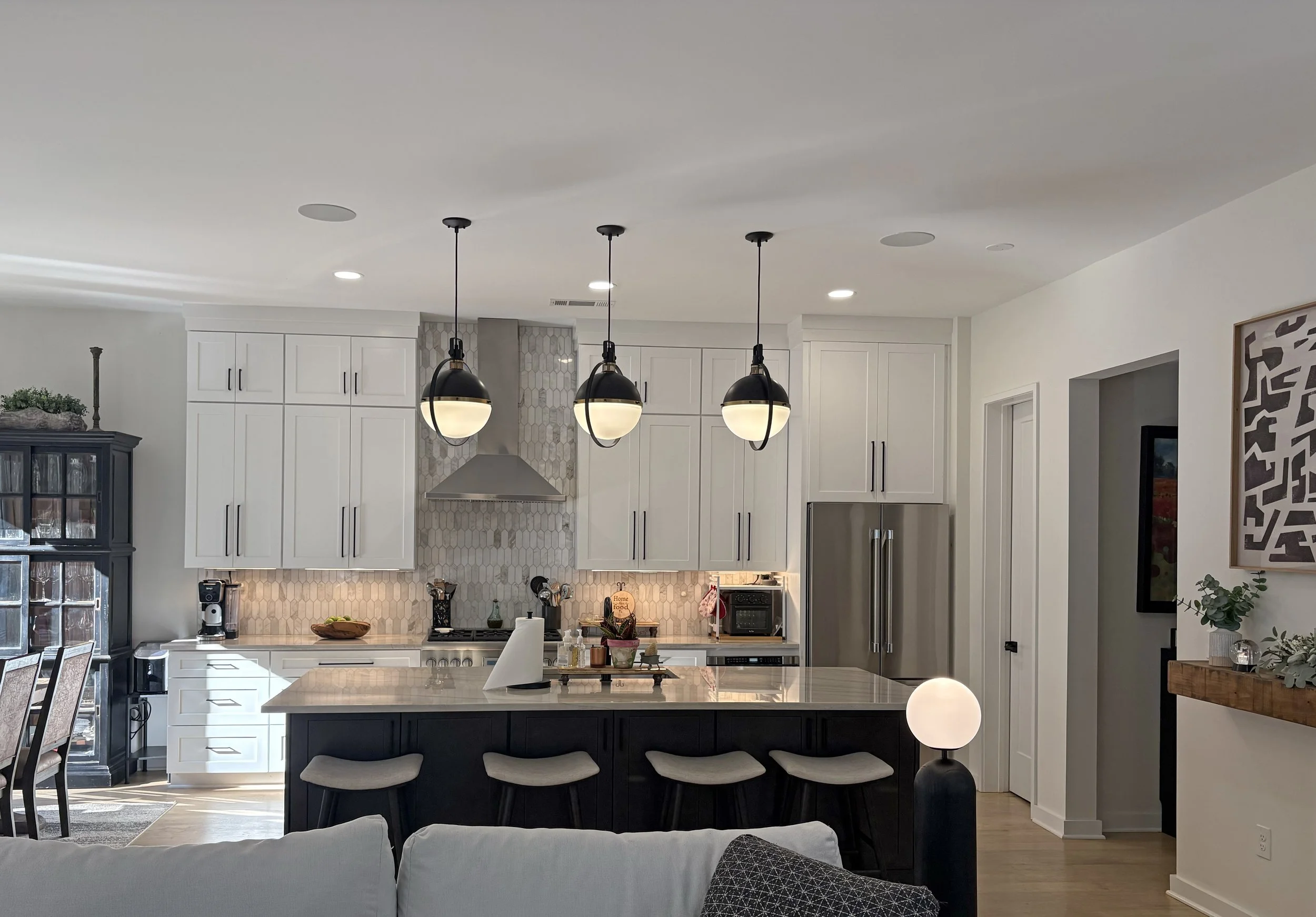 Modern kitchen with white cabinets, marble island, three black pendant lights, stainless steel refrigerator, and a built-in oven, with decor and appliances on the countertop.