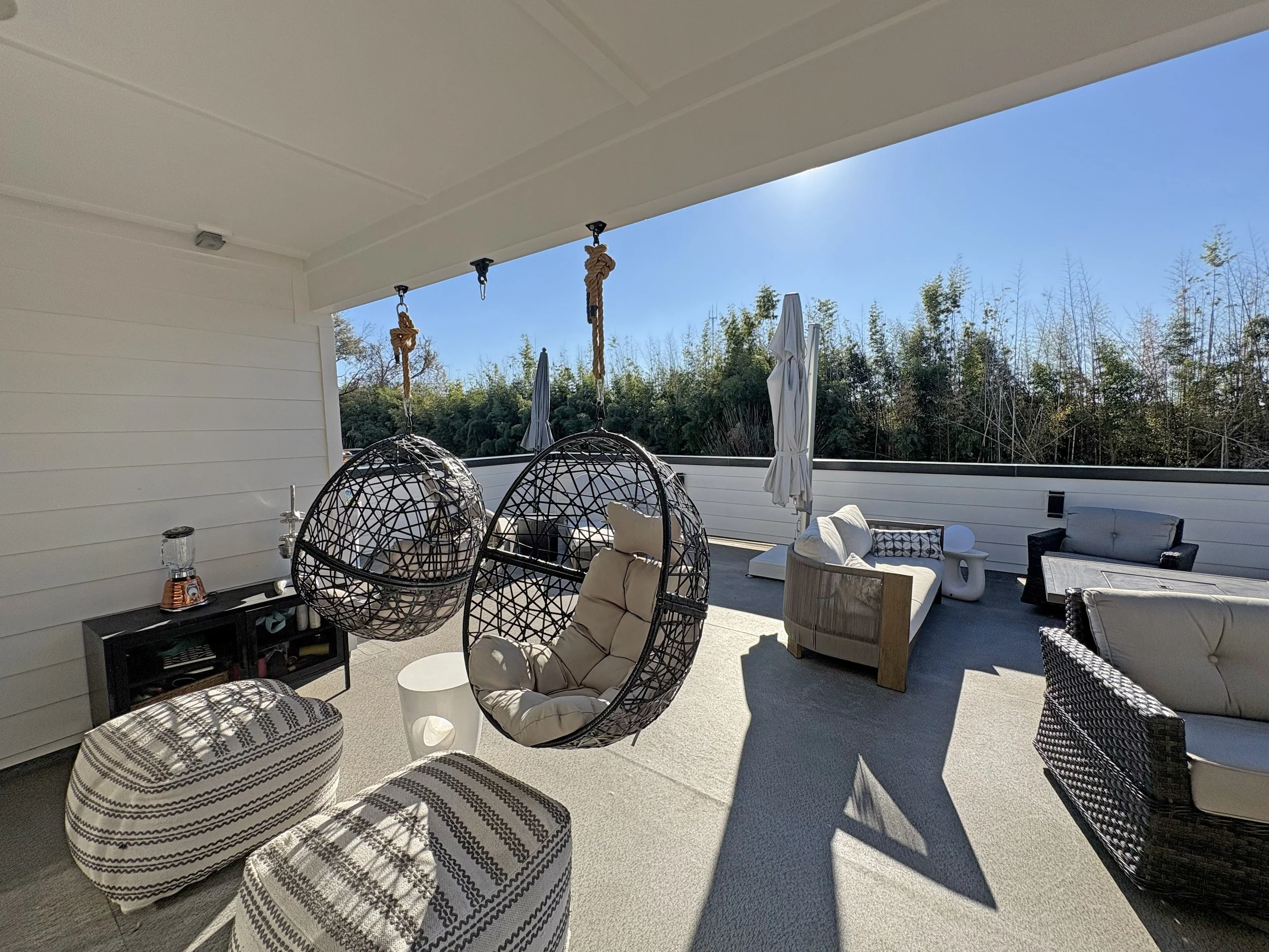 Covered outdoor patio with hanging black woven egg chairs with cushions, seating area with sofas and chairs, a small table, and closed patio umbrellas, all with a view of trees under a clear blue sky.