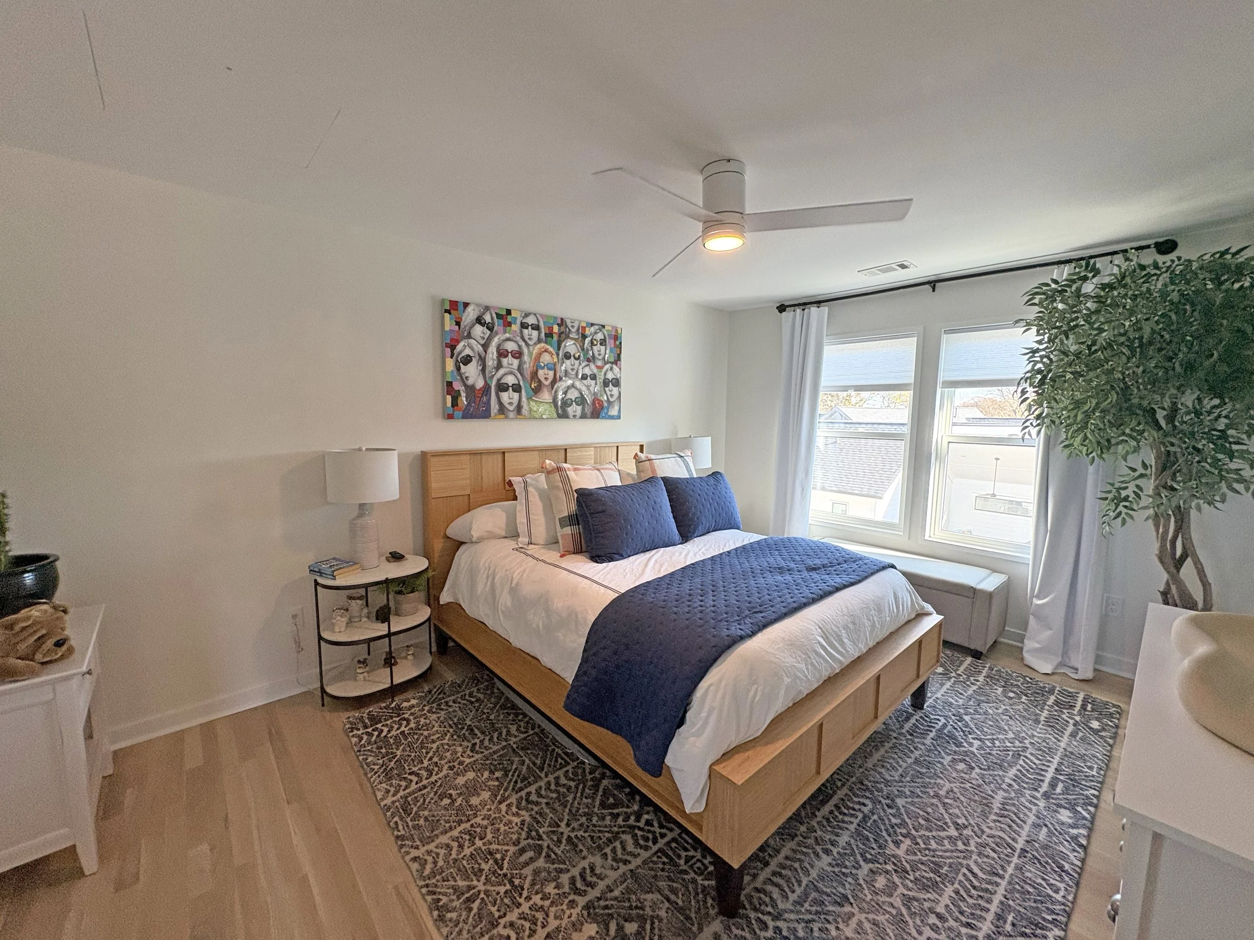 A modern bedroom with a wooden bed frame, white bedding, and navy blue pillows and throw blanket. There is a white nightstand with a lamp, books, and decor. A colorful artwork of women with sunglasses hangs on the white wall above the bed. Large wind
