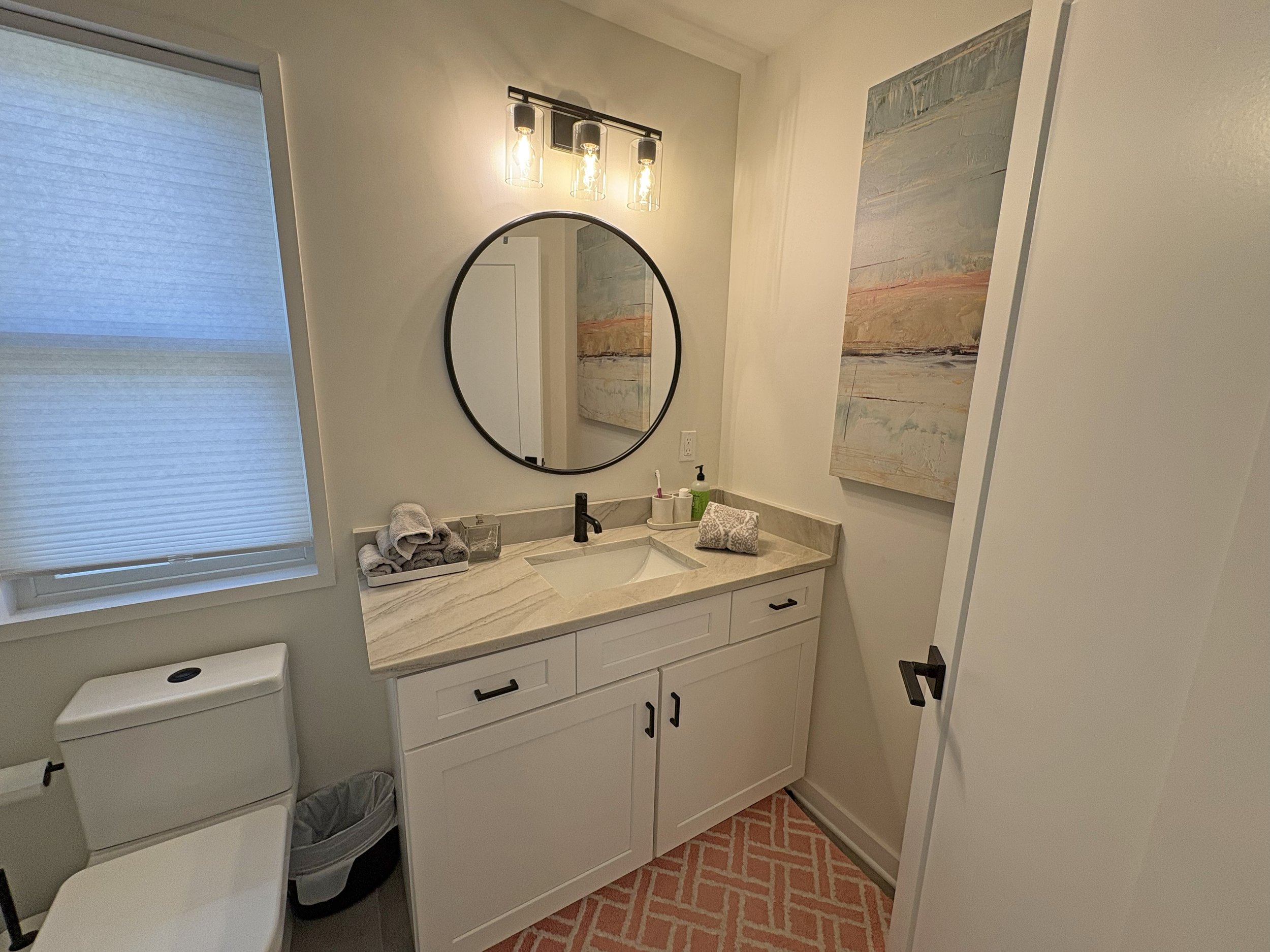 Bathroom with white vanity, marble countertop, round mirror, black faucet, towels, toiletries, and a large abstract painting. There's a window with a light-colored shade, a toilet, and a patterned pink rug on the floor.