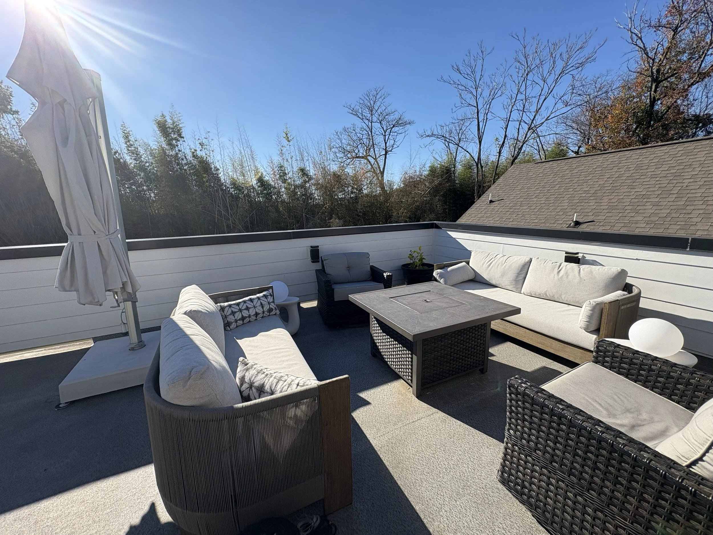 Outdoor patio with cream and black wicker furniture, cushions, a table, and a closed umbrella, surrounded by a white fence and trees with a clear blue sky.