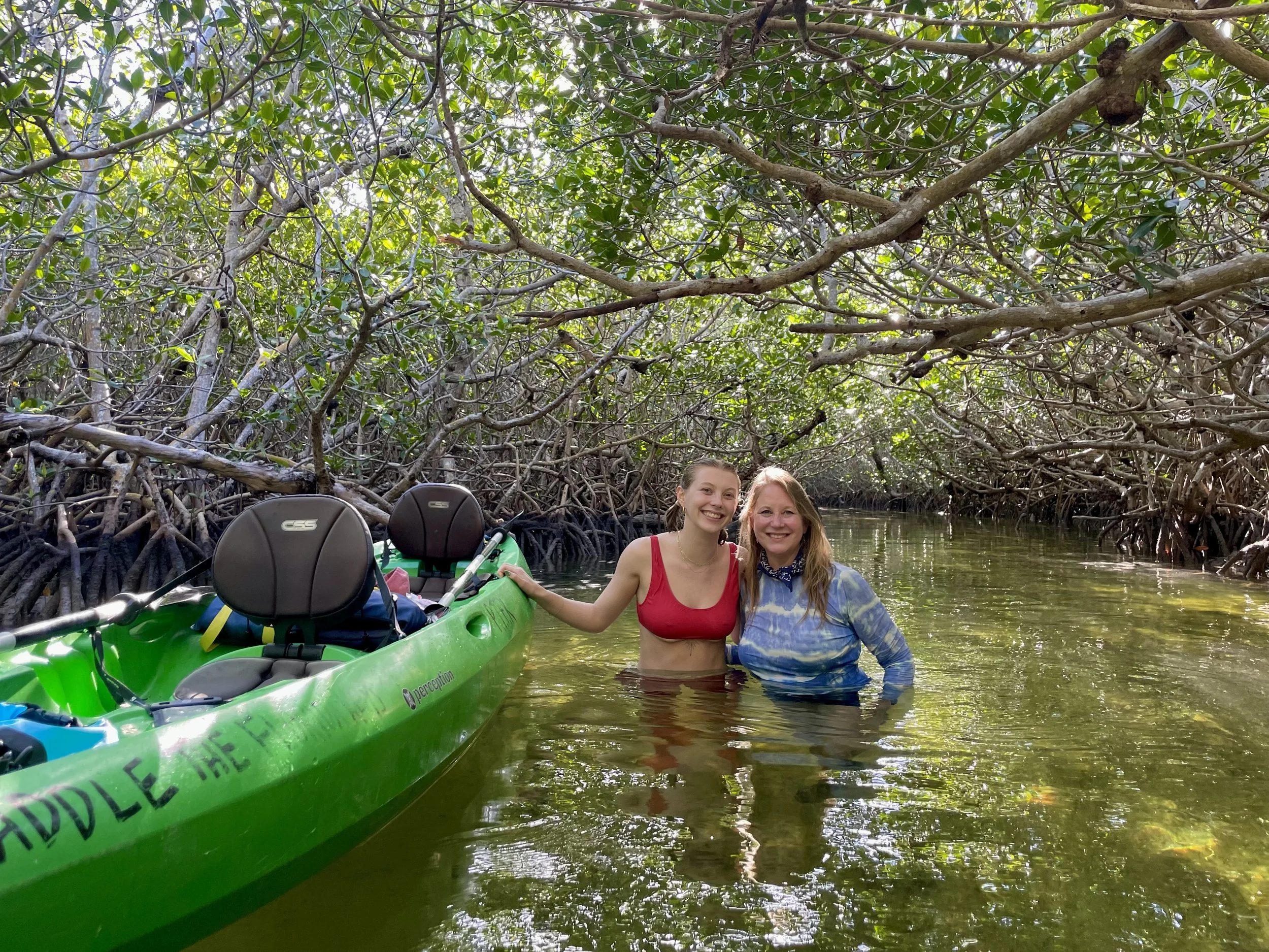Two women standing in a narrow waterway surrounded by dense mangrove trees, with a lime green kayak nearby.