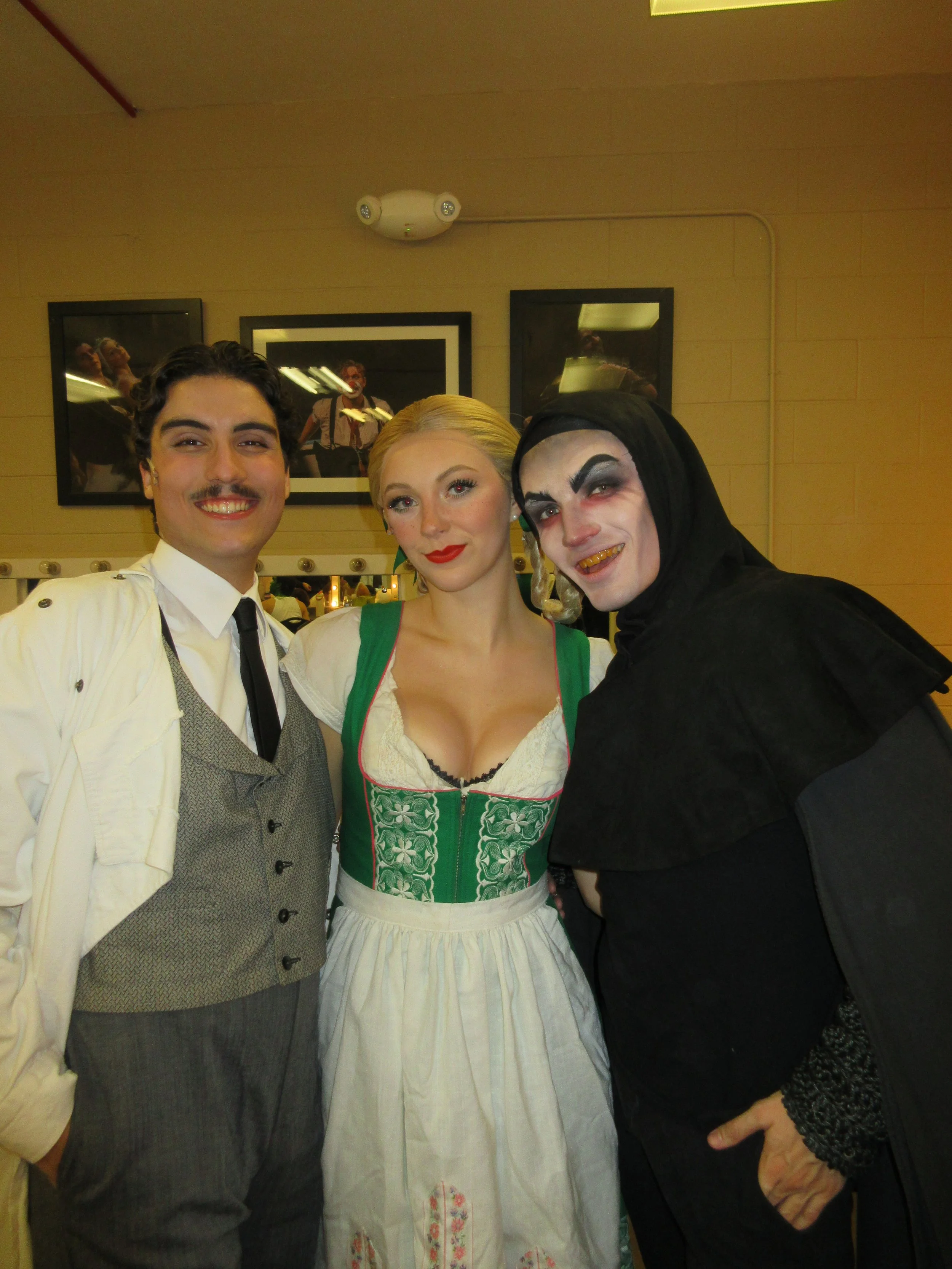 Three people in costume posing together, with two framed pictures and a security camera on the wall behind them.