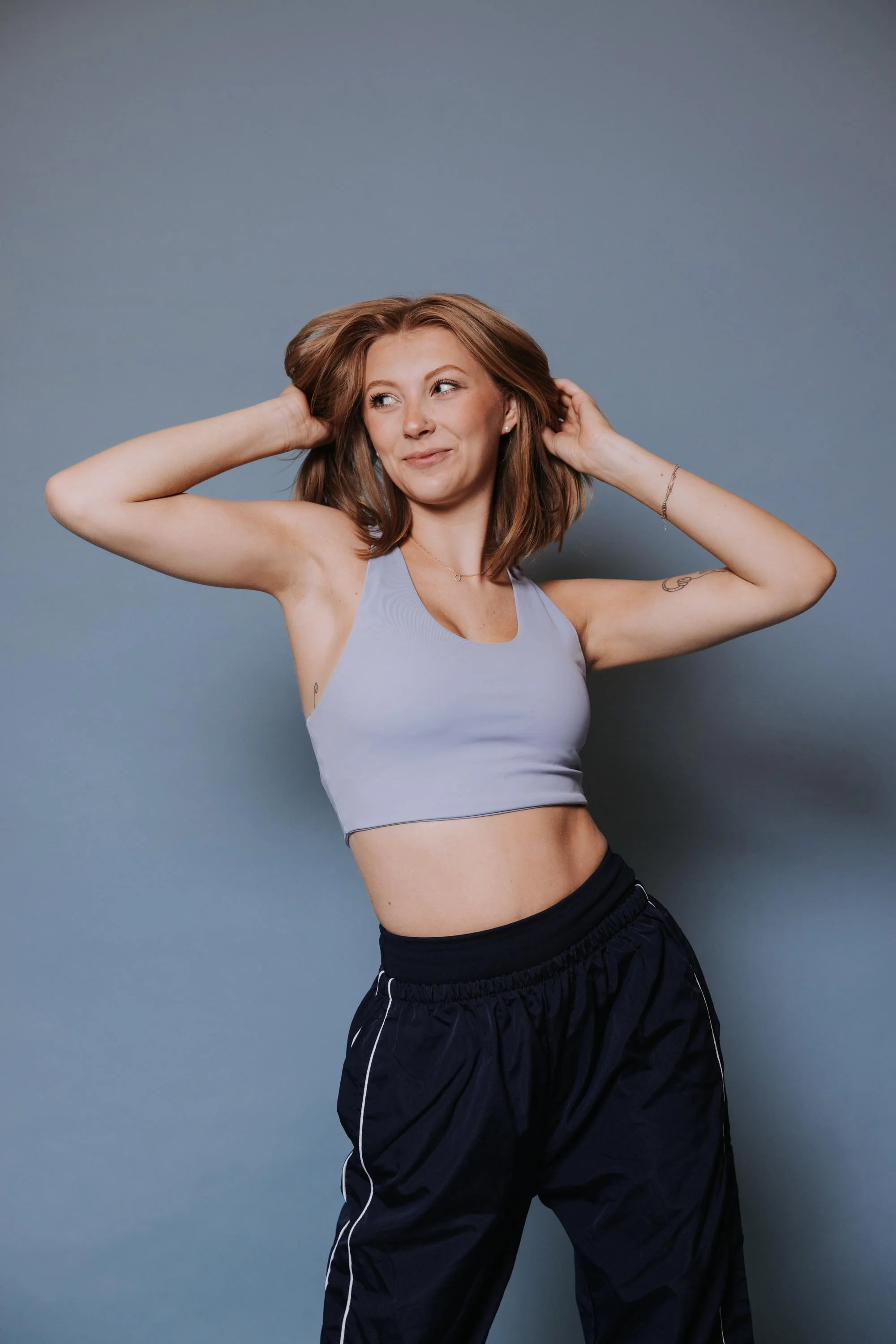 A young woman with shoulder-length brown hair and light skin, wearing a light purple sports bra and black athletic pants, standing against a gray background and posing with her hands behind her head.