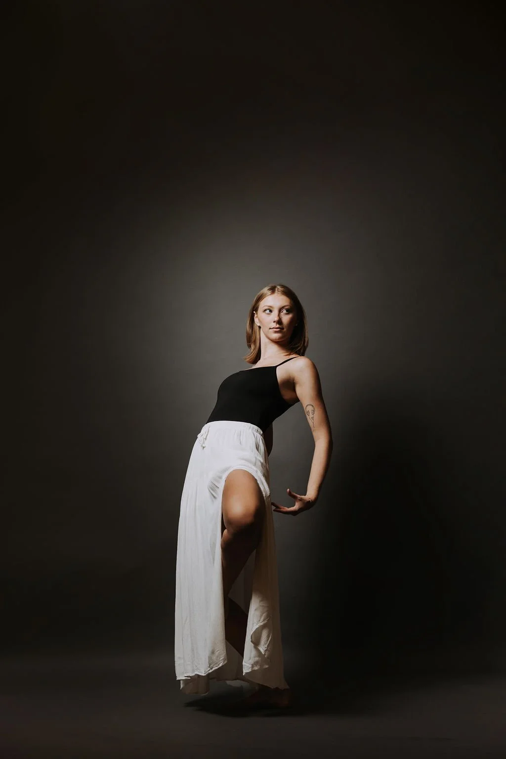 A young woman with shoulder-length brown hair wearing a black tank top and white wide-leg pants, standing against a dark background, posing with one hand on her hip and the other arm bent, looking to her side.