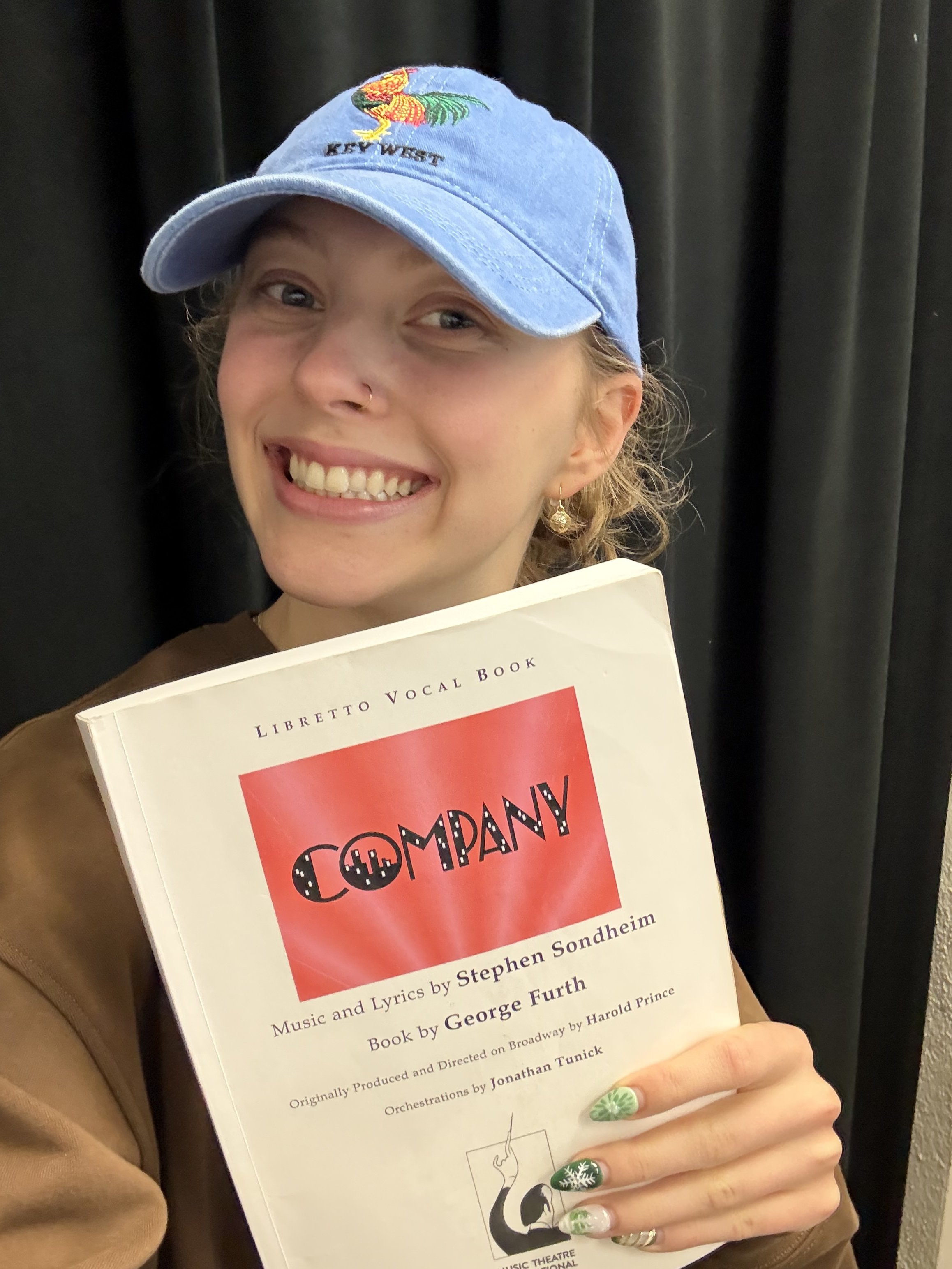 A woman smiling while holding a musical theater vocal book titled "Company" by Stephen Sondheim, wearing a blue baseball cap and a brown top.