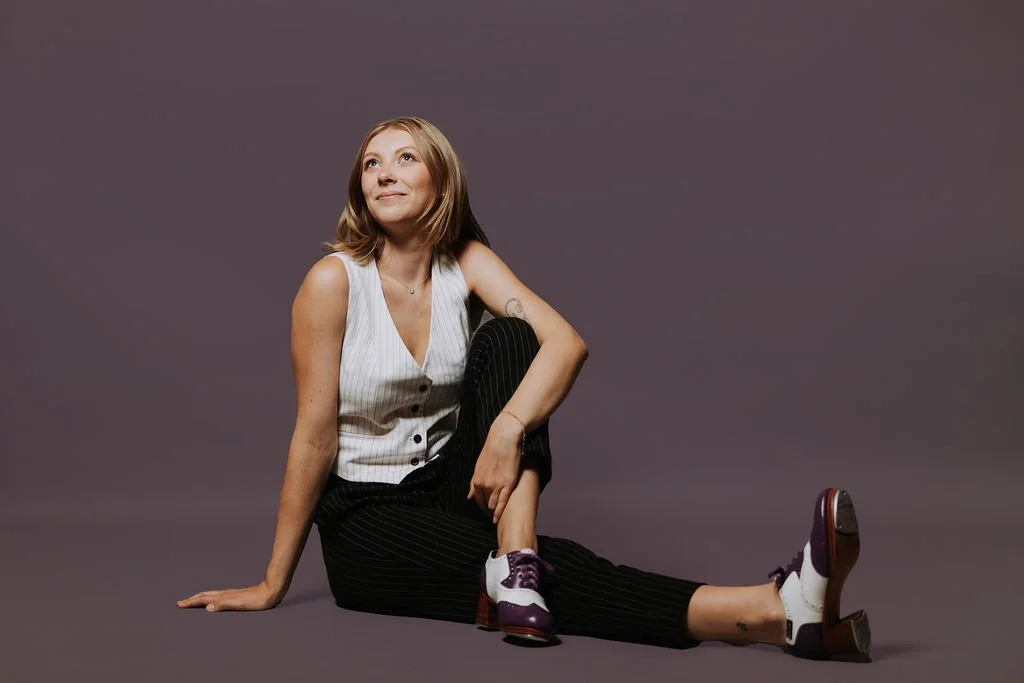 A young woman with blonde hair sitting on the floor against a plain gray background, wearing a sleeveless white top, black pinstripe pants, and white and purple shoes, looking upwards with a thoughtful expression.