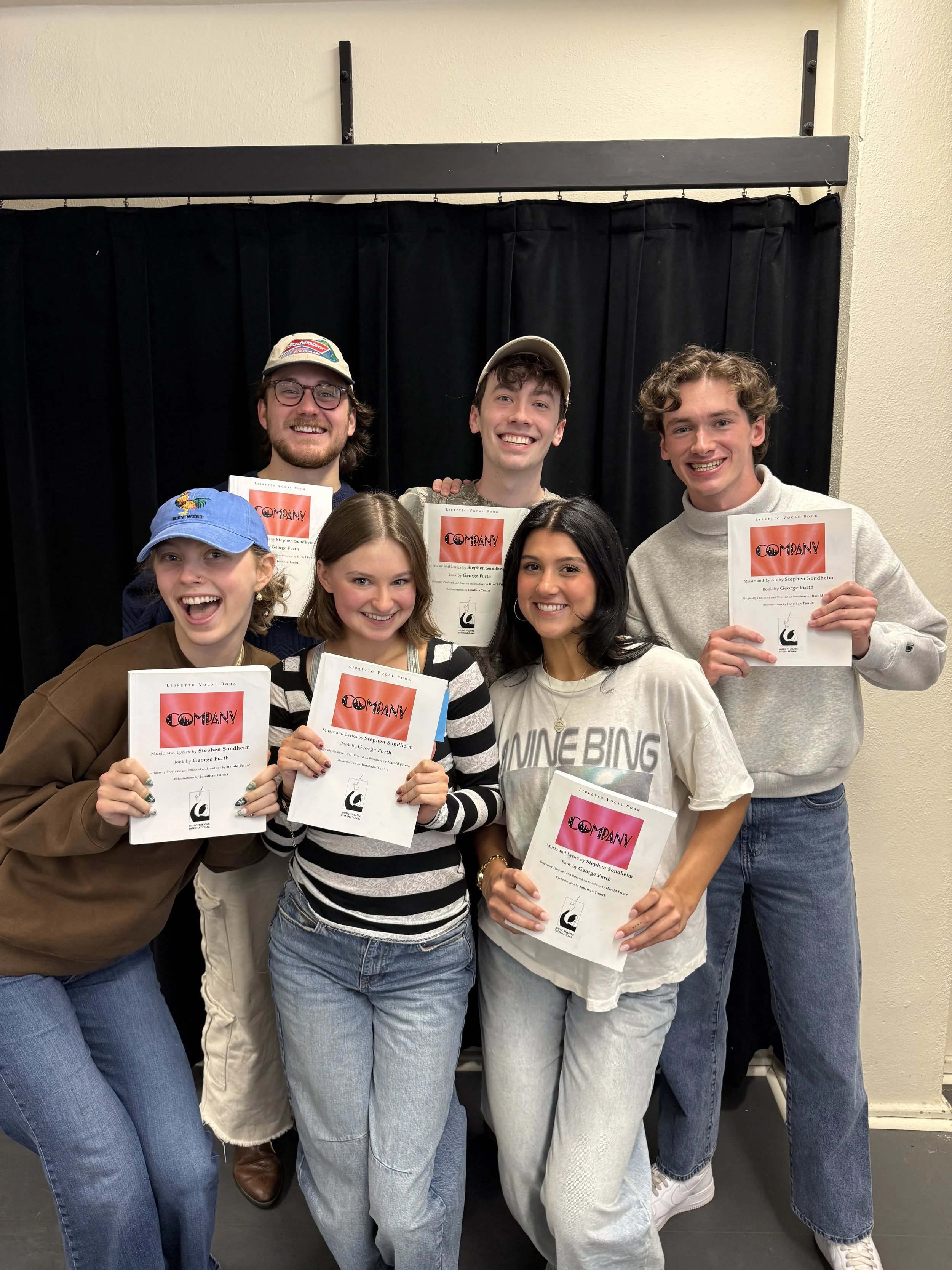Seven young adults smiling and holding sheet music and certificates, standing in front of a black curtain backdrop, celebrating a musical achievement.