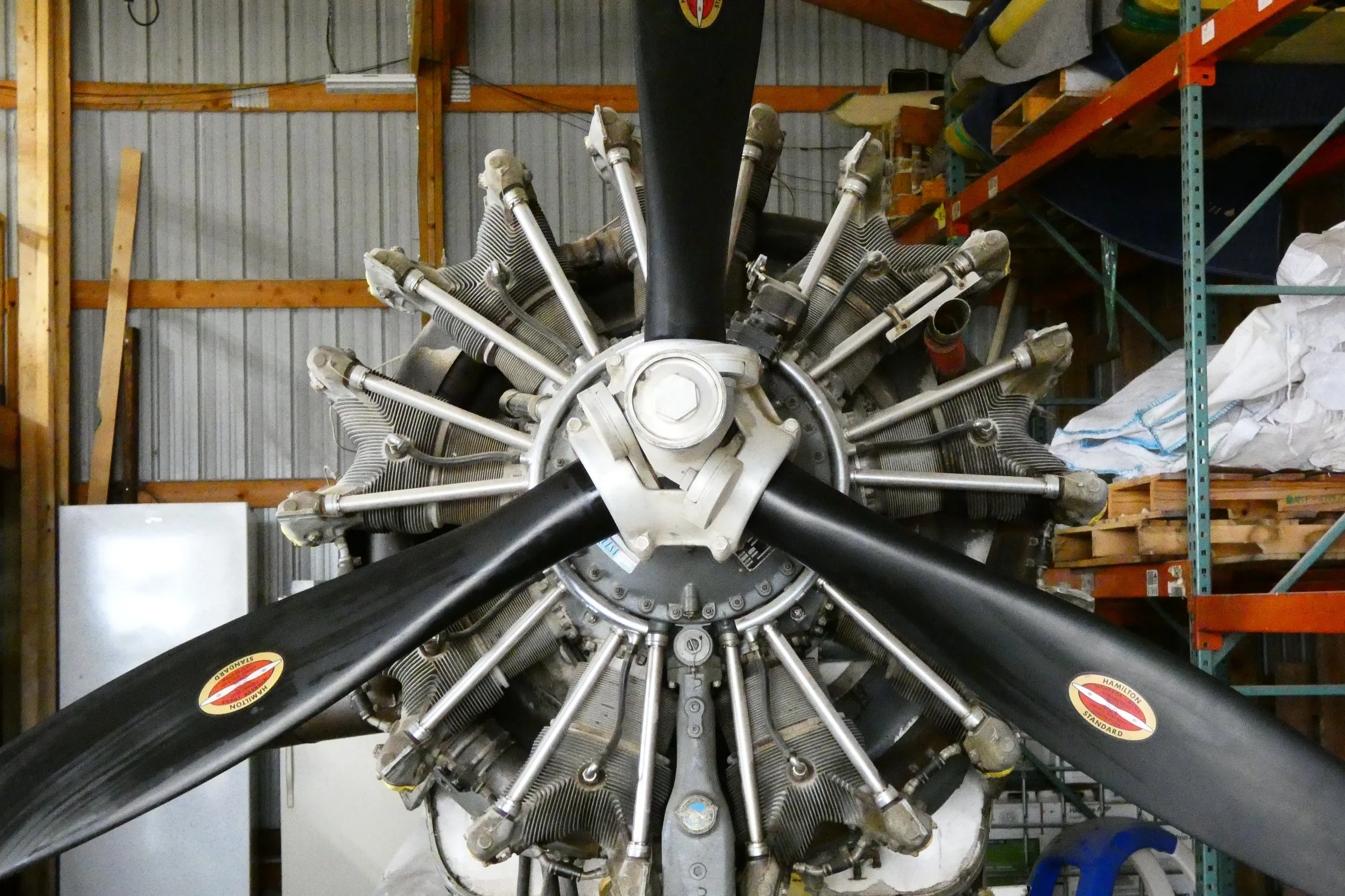 Front view of a vintage radial aircraft engine with a black propeller attached, situated inside a storage shed.