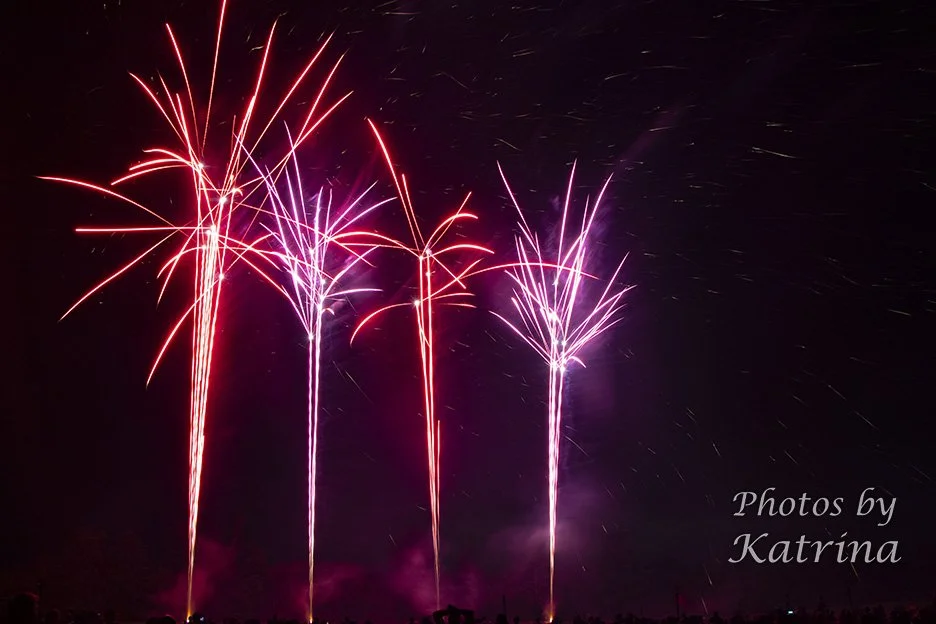 Fireworks exploding in pink and red colors in the night sky, with a credit to photos by Katrina.