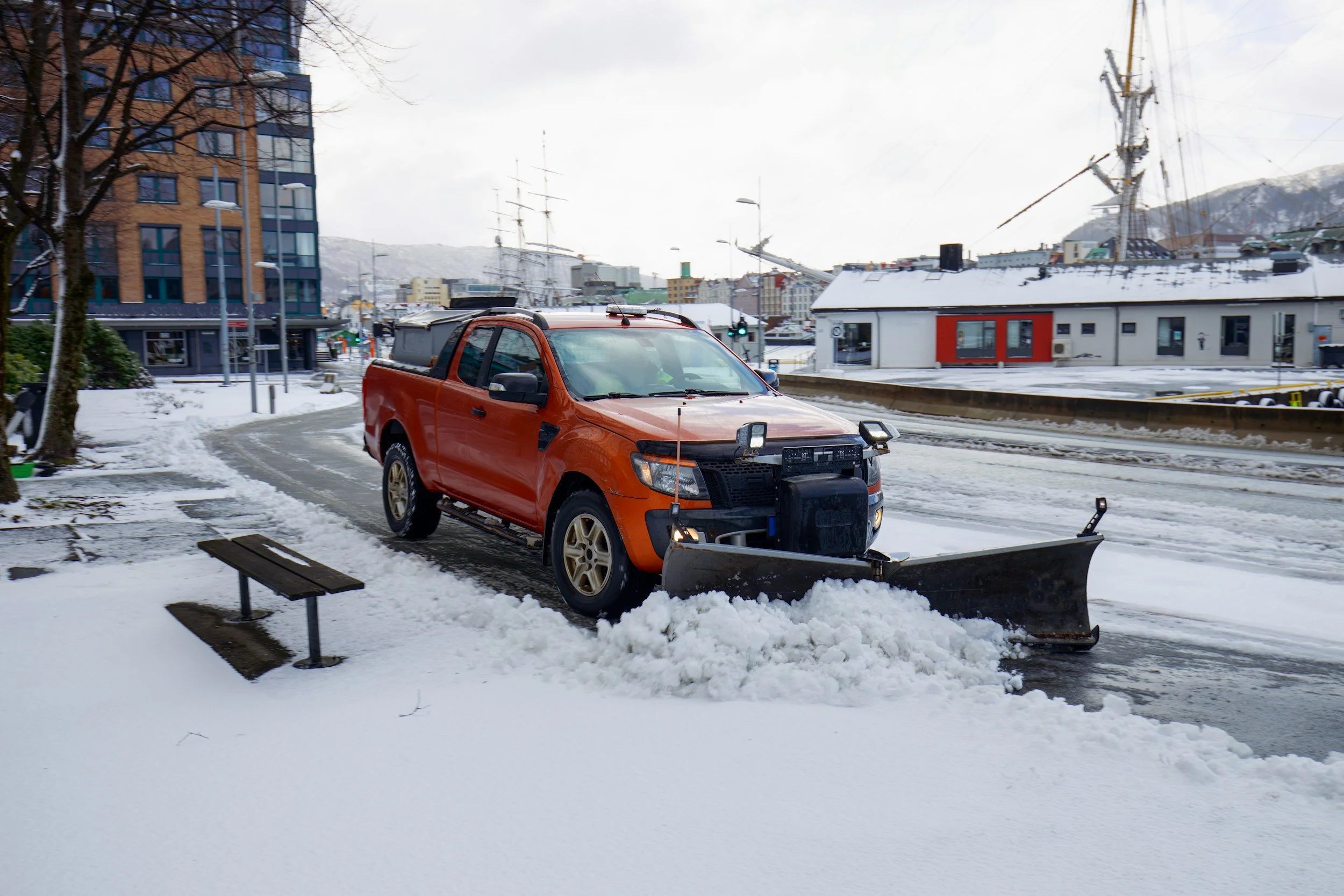Orange pickup truck equipped with a snow plow clearing snow from a city street.