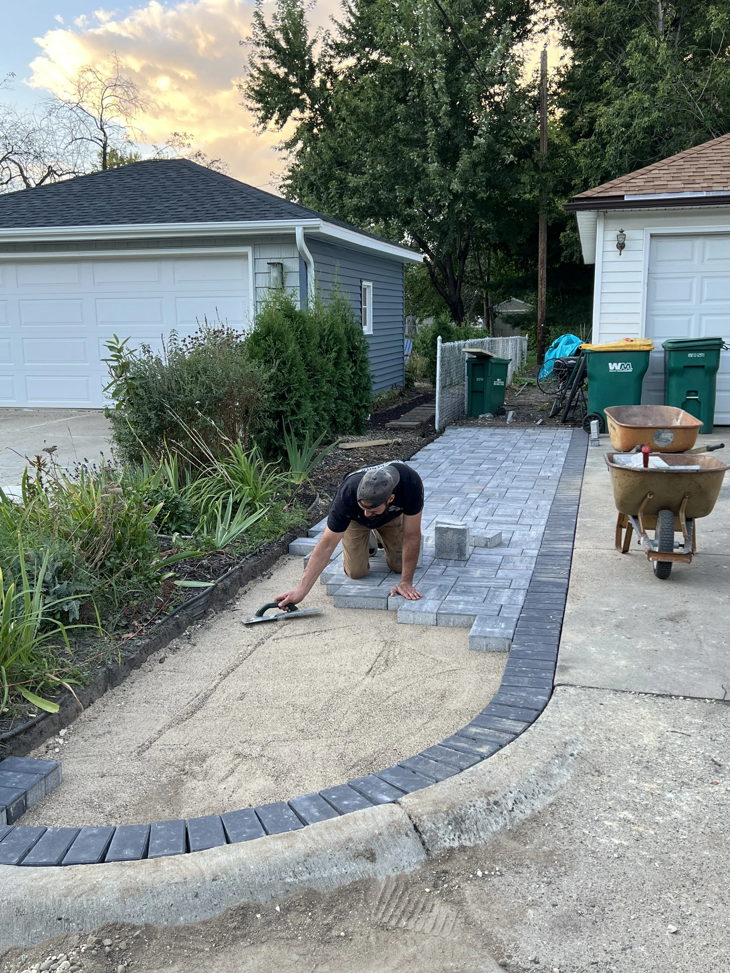 A person laying paving stone bricks to create a curved pathway in a backyard during sunset, with tools and materials around.