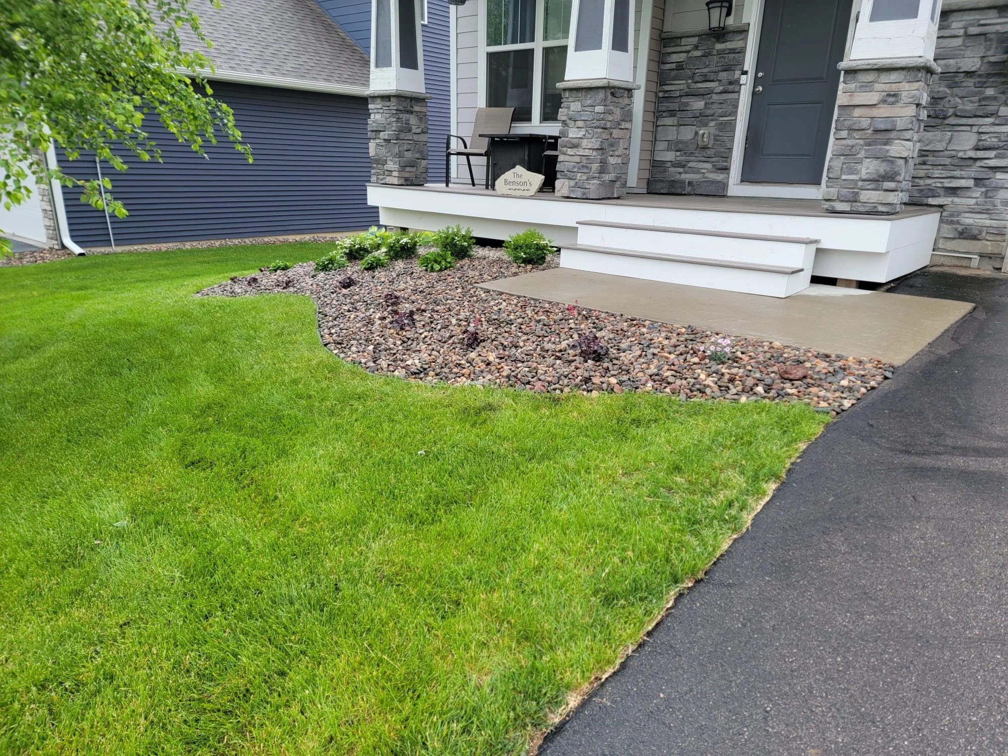 Front yard with green grass, small shrubbery, and a walkway leading to a porch with stairs, surrounded by stone and gravel landscaping.