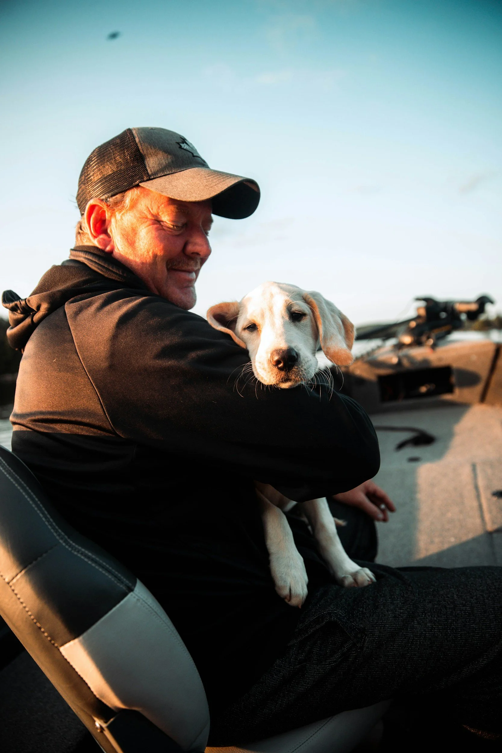 A man wearing a baseball cap and black jacket is sitting in a vehicle, holding a young light-colored puppy with floppy ears. The man is smiling and the puppy appears calm. The background shows a clear blue sky and part of a boat or vehicle.